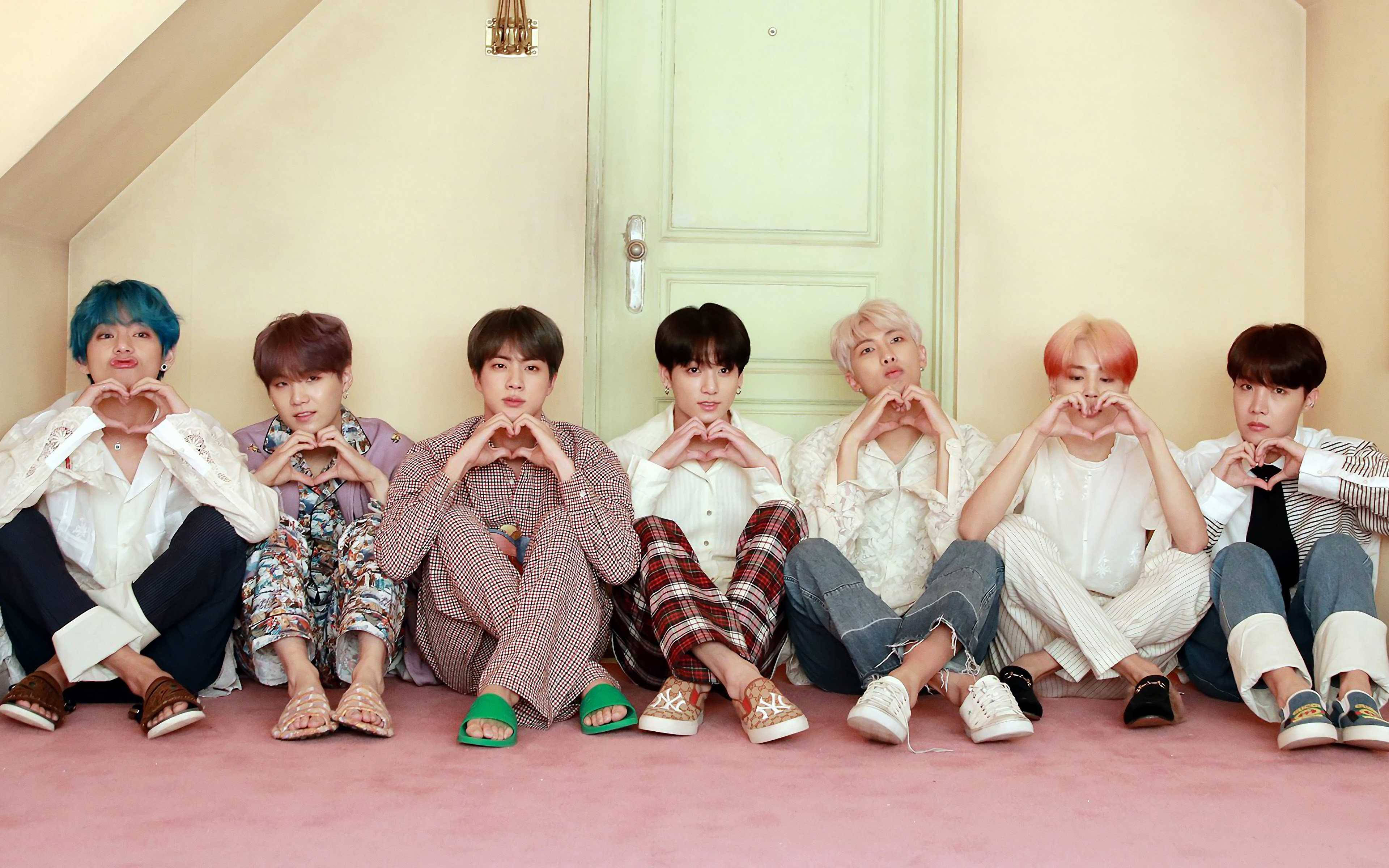 BTS 桌布 桌布 壁紙 — 七位年輕男子擁有色彩繽紛的頭髮，坐在粉紅色地板上，對著一扇粉綠色門用手做出心形。