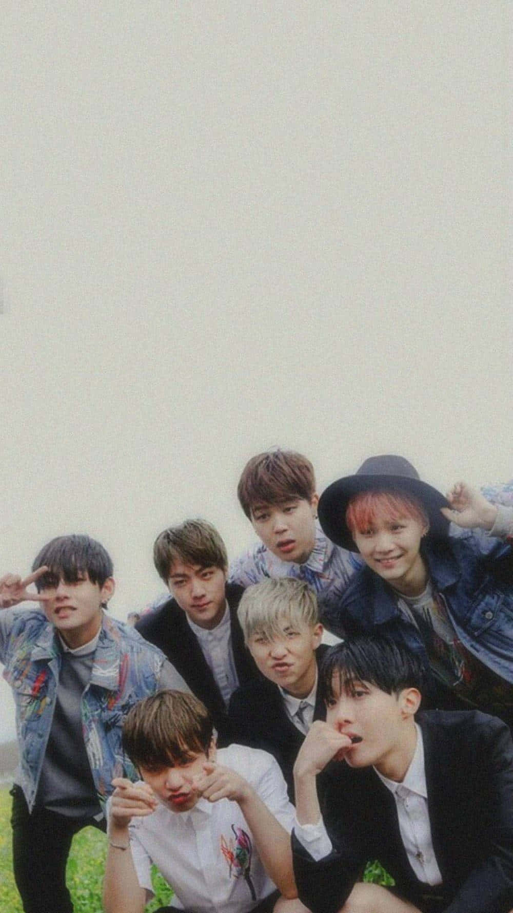 BTS 桌布 壁紙 — 七位年輕男子身穿休閒服裝，在草地上以多種表情和手勢，對著朦朧的天空擺出俏皮的姿勢。