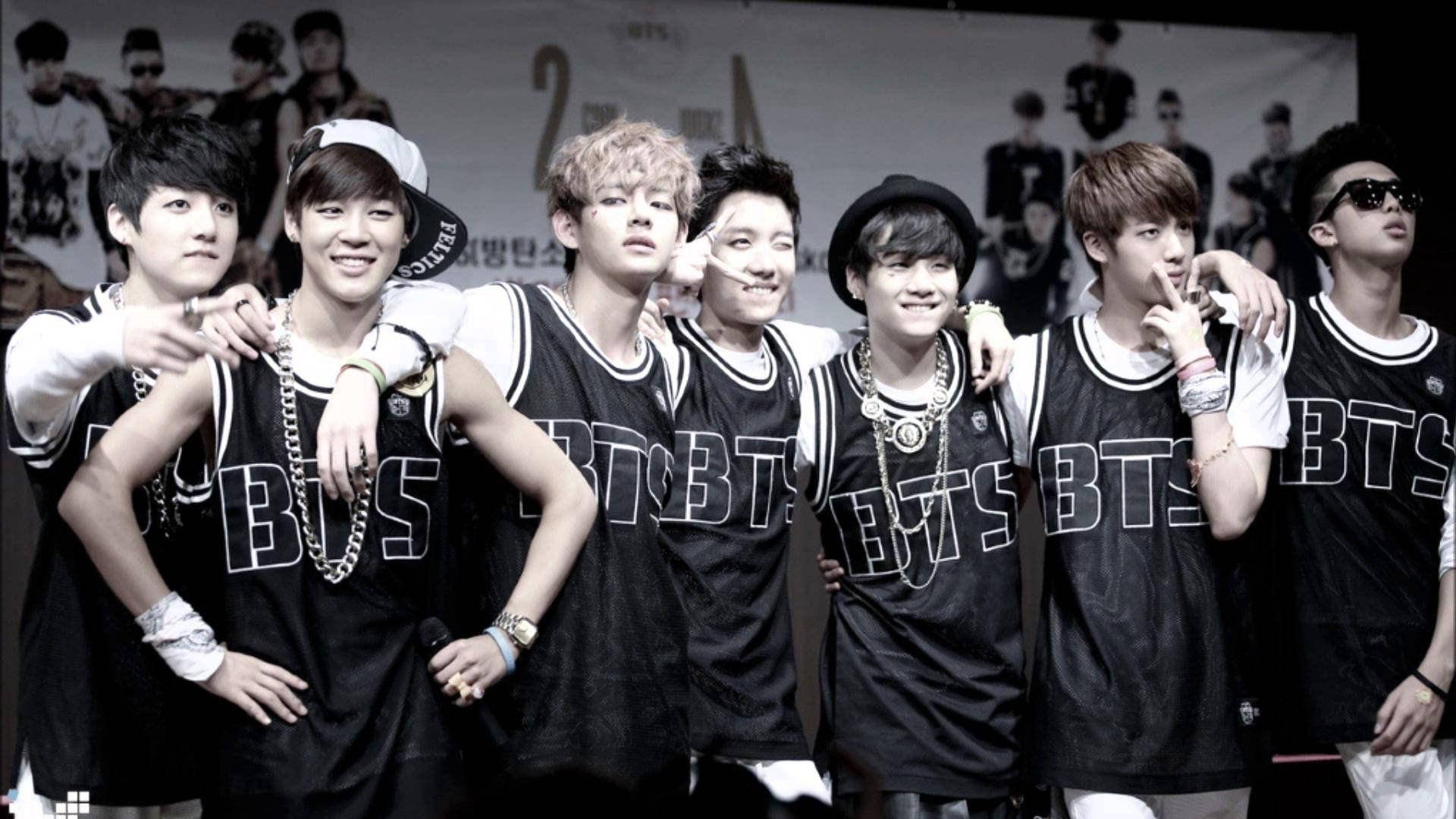 BTS 2021 桌布 壁紙 — 七位年輕男子身穿相配的黑色「BTS」球衣，在宣傳背景前自信地擺出姿勢，搭配不同飾品和表情。