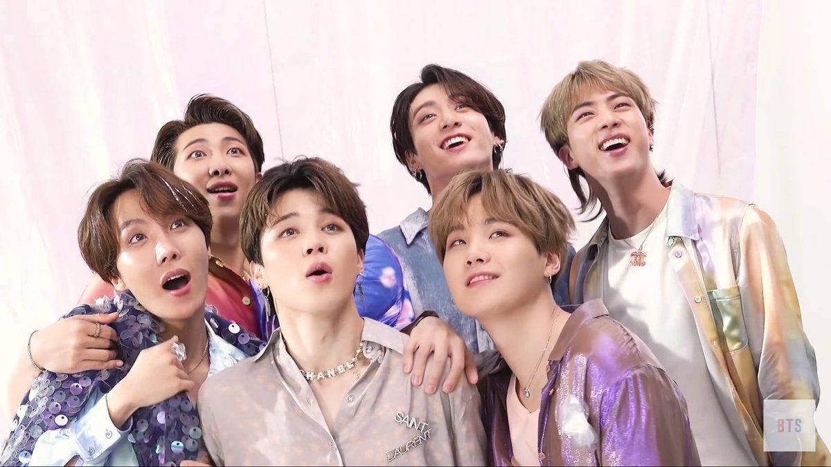 BTS 桌布 壁紙 — 七位年輕男子身穿閃亮多彩的服裝，在柔和的粉紅色窗簾背景下，帶著歡樂的表情俏皮地擺姿勢。