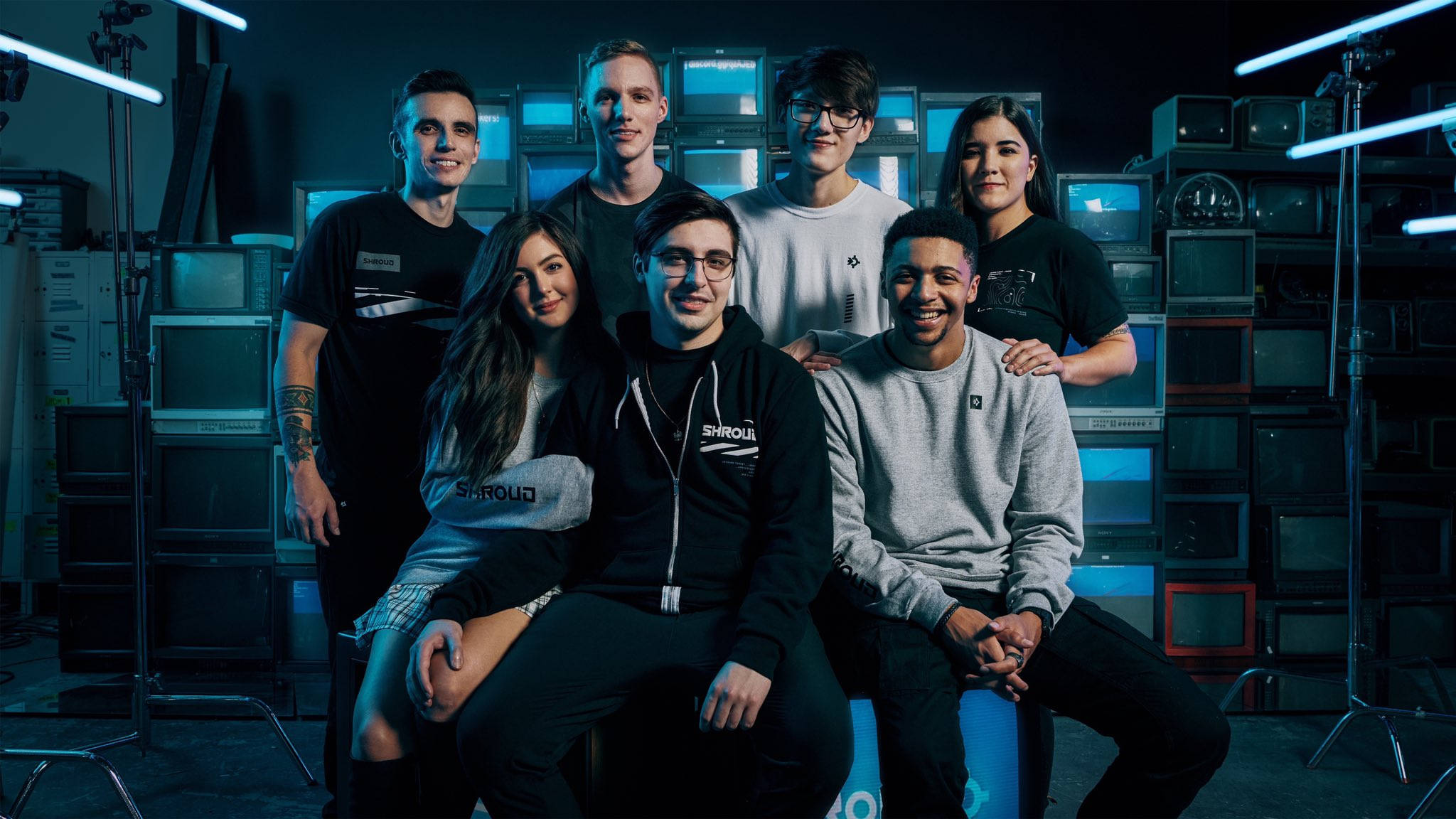 Shroud 桌布 壁紙 — 七位微笑的年輕成年人在一個黑暗的攝影棚中自信地擺姿勢，周圍環繞著發出藍光的復古電視機。