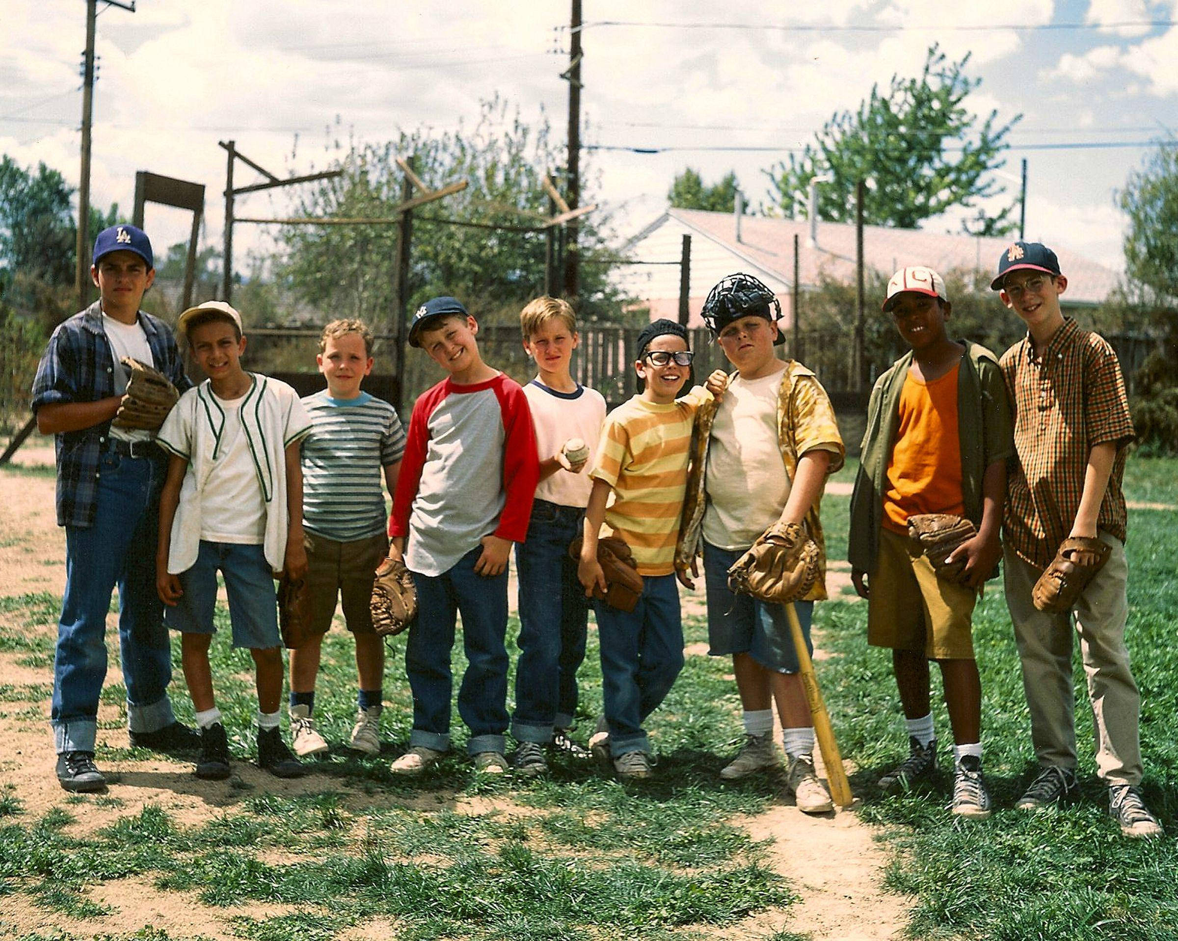 The Sandlot 桌布 壁紙 — 七位微笑的年輕棒球運動員穿著復古制服和帽子，在陽光明媚的草地上自豪地擺姿勢，手持手套和球棒。