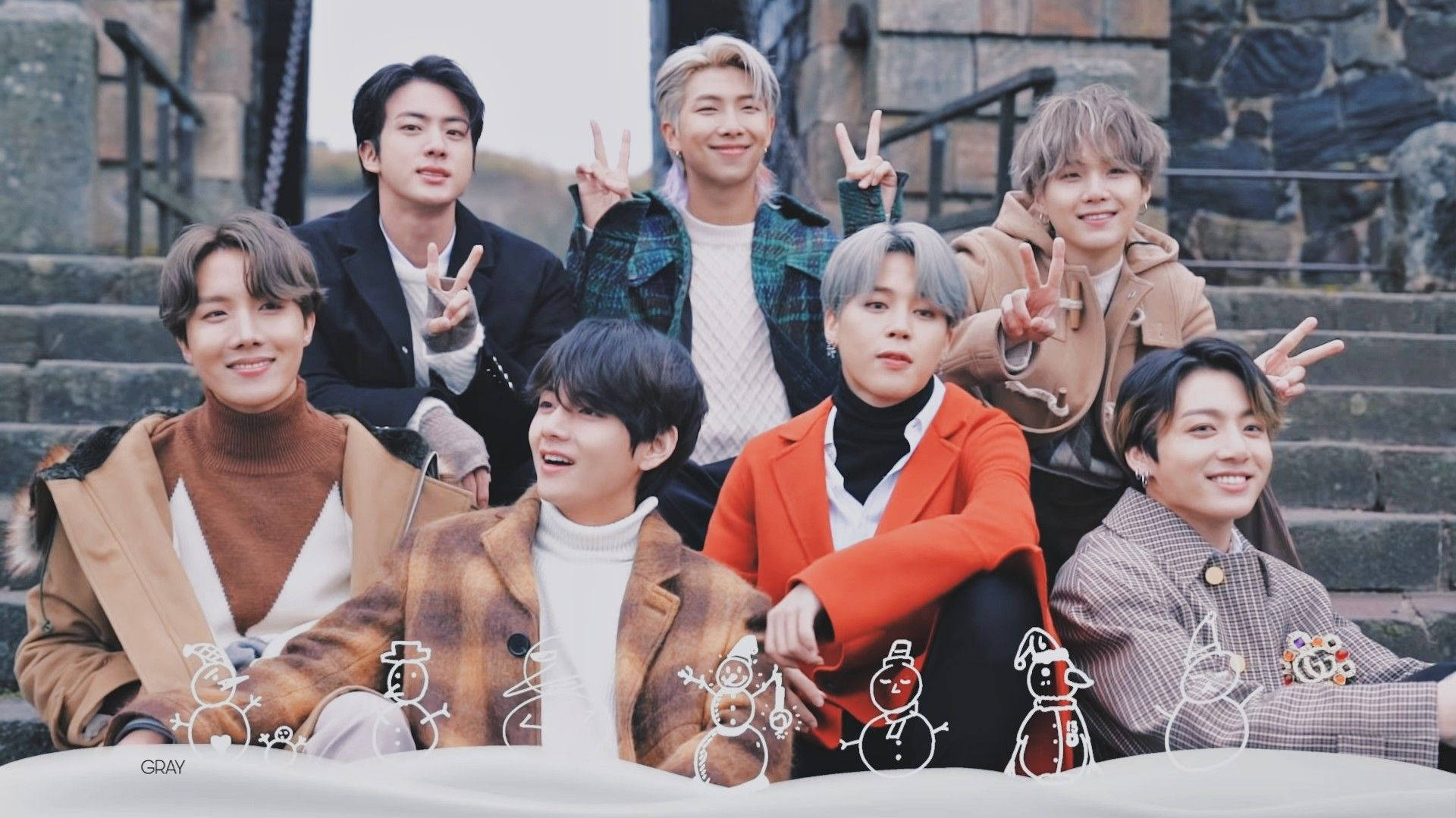 BTS 桌布 桌布 壁紙 — 七位微笑的年輕男士穿著時尚冬季外套，在石階上愉快地擺姿勢，底部有雪人塗鴉。