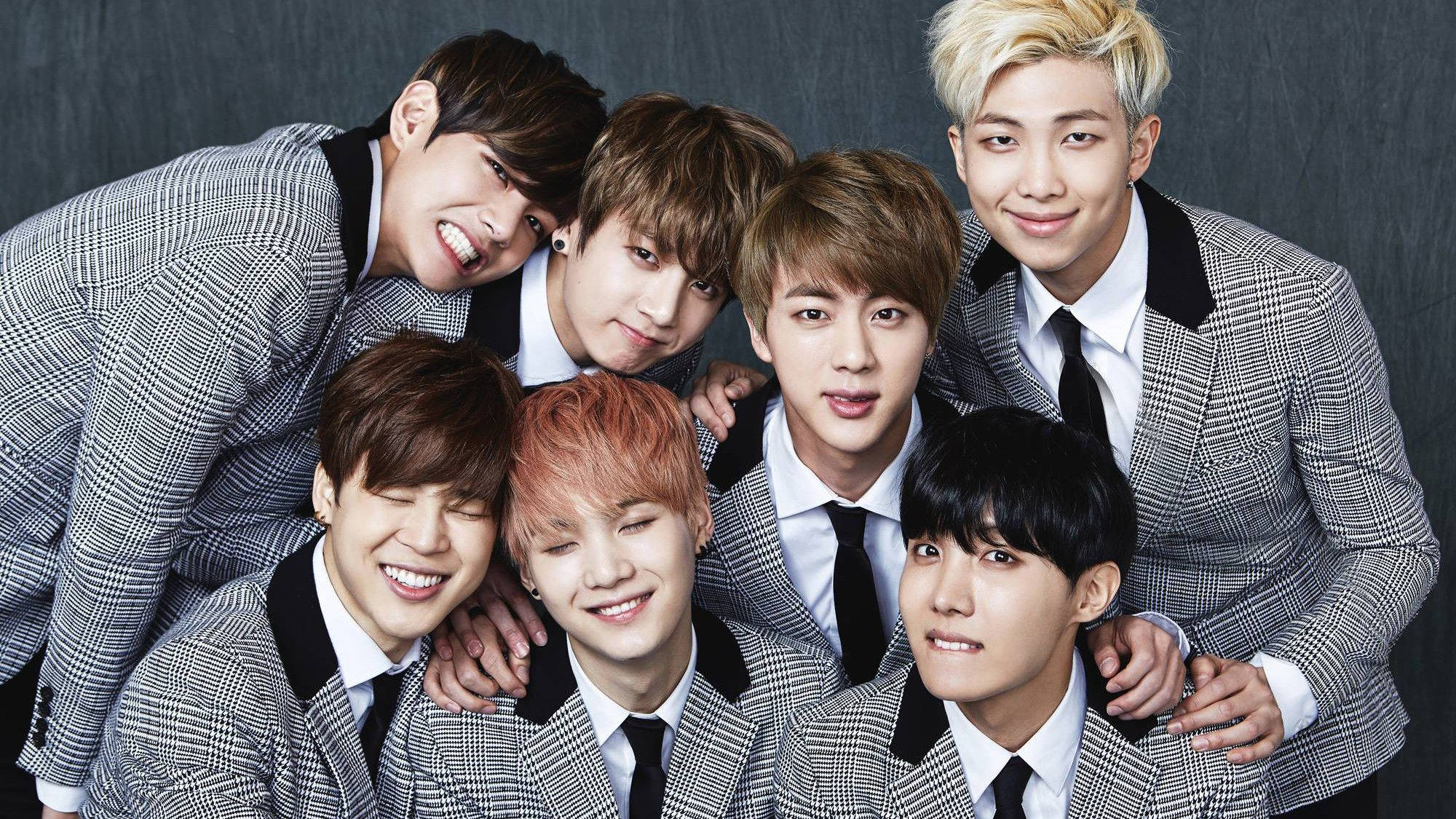 BTS 桌布 壁紙 — 七位微笑的年輕男士穿著相配的灰色格紋西裝，在深色紋理背景前緊密地擺姿勢。