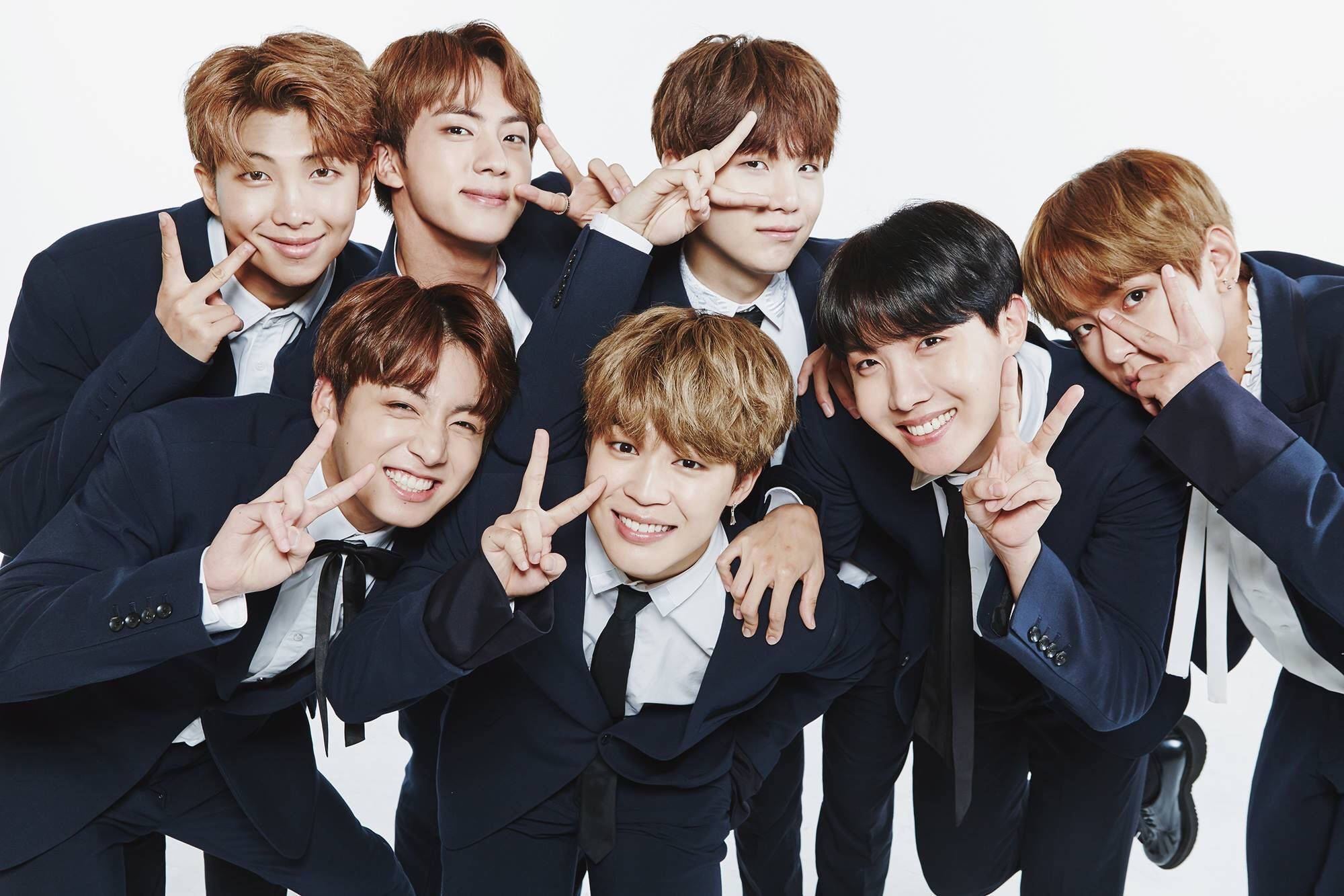 BTS 桌布 壁紙 — 七位微笑的年輕男士身穿相配的海軍藍西裝，在純白背景前緊密地擺出和平手勢。