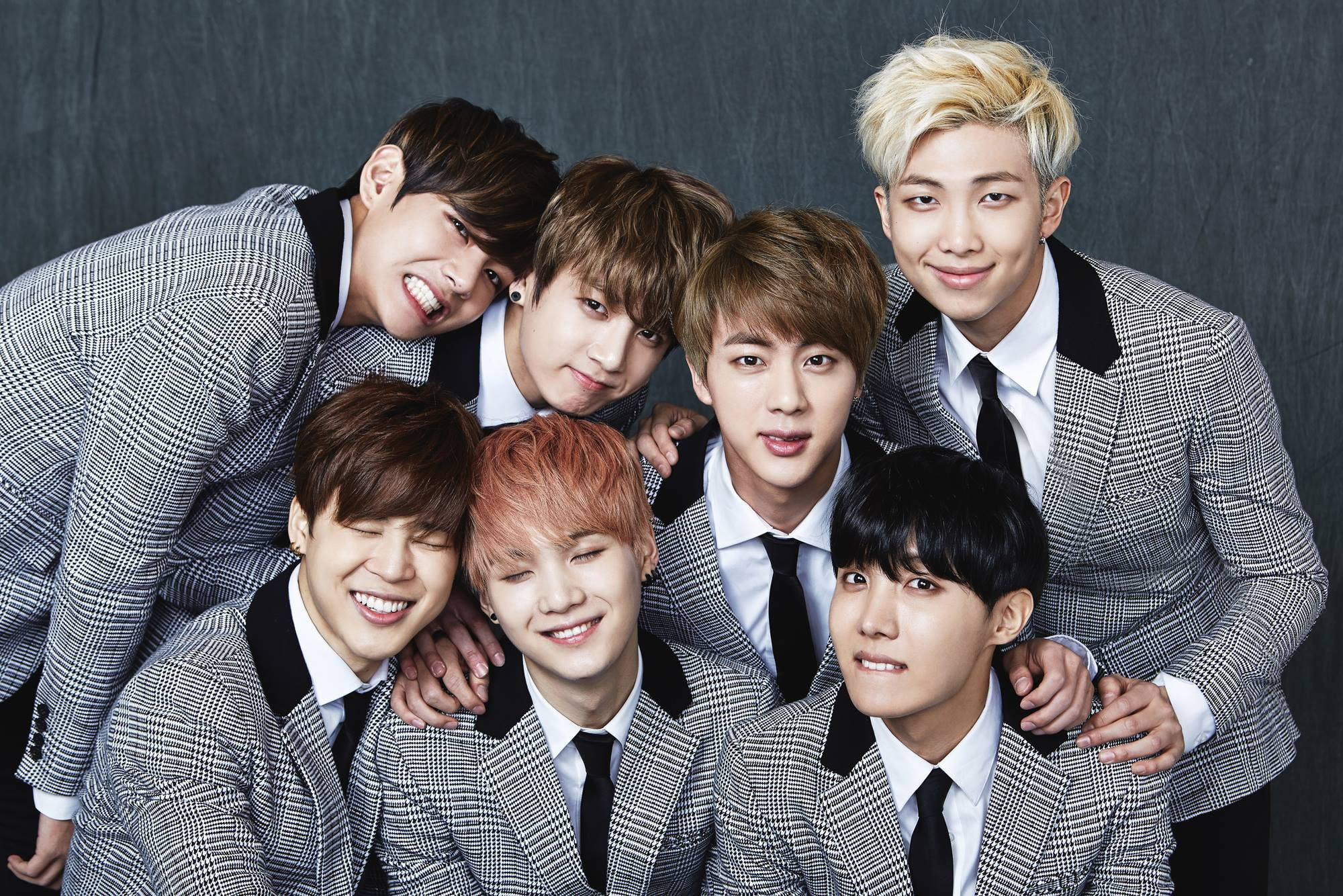 BTS 桌布 壁紙 — 七位微笑的年輕男士身穿相配的灰色格紋西裝，在深色紋理背景前緊密地擺姿勢。