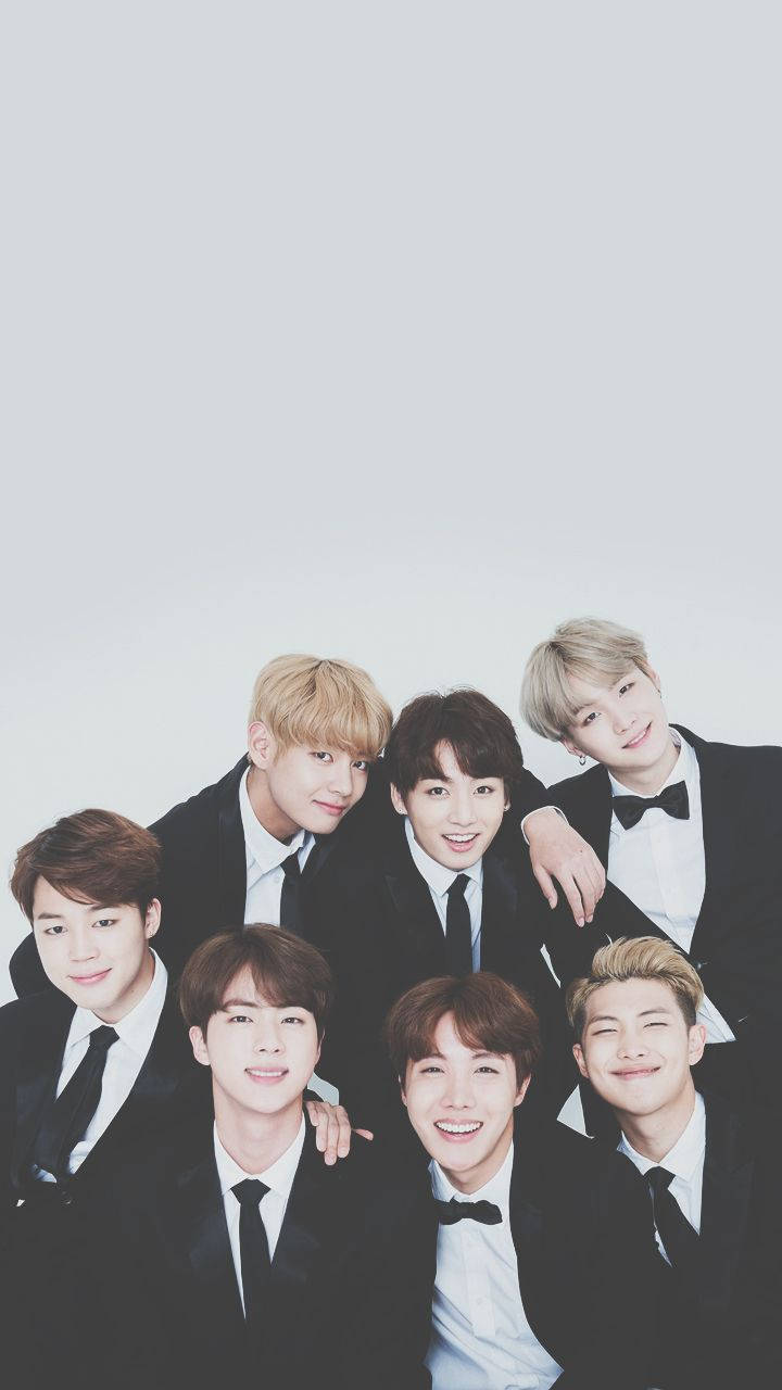 BTS 桌布 壁紙 — 七位微笑的年輕男士身穿黑色燕尾服，在淺灰色背景前緊密地擺姿勢。