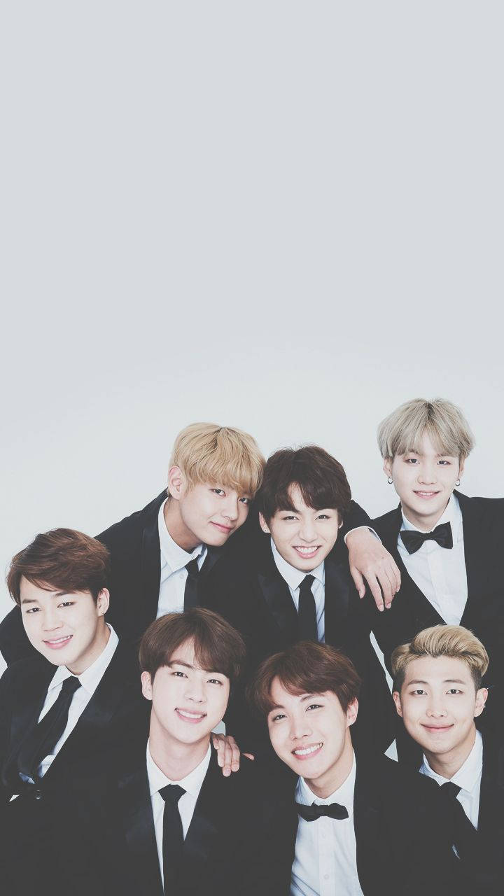 BTS 桌布 壁紙 — 七位微笑的年輕男士身穿黑色禮服，緊密地站在純淺灰色背景前擺姿勢。