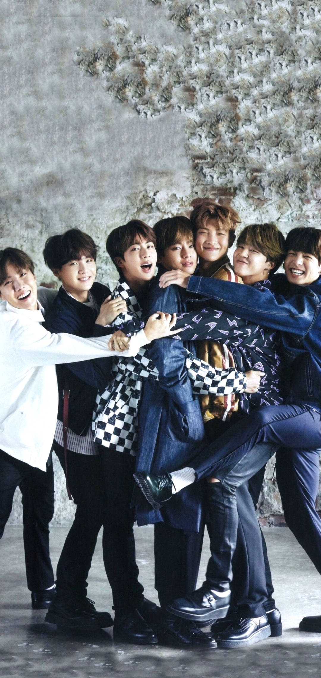 BTS 鎖定螢幕 桌布 壁紙 — 七位微笑的年輕男子穿著休閒服裝，擠在一起靠著紋理混凝土牆，擺出歡樂的團體姿勢。