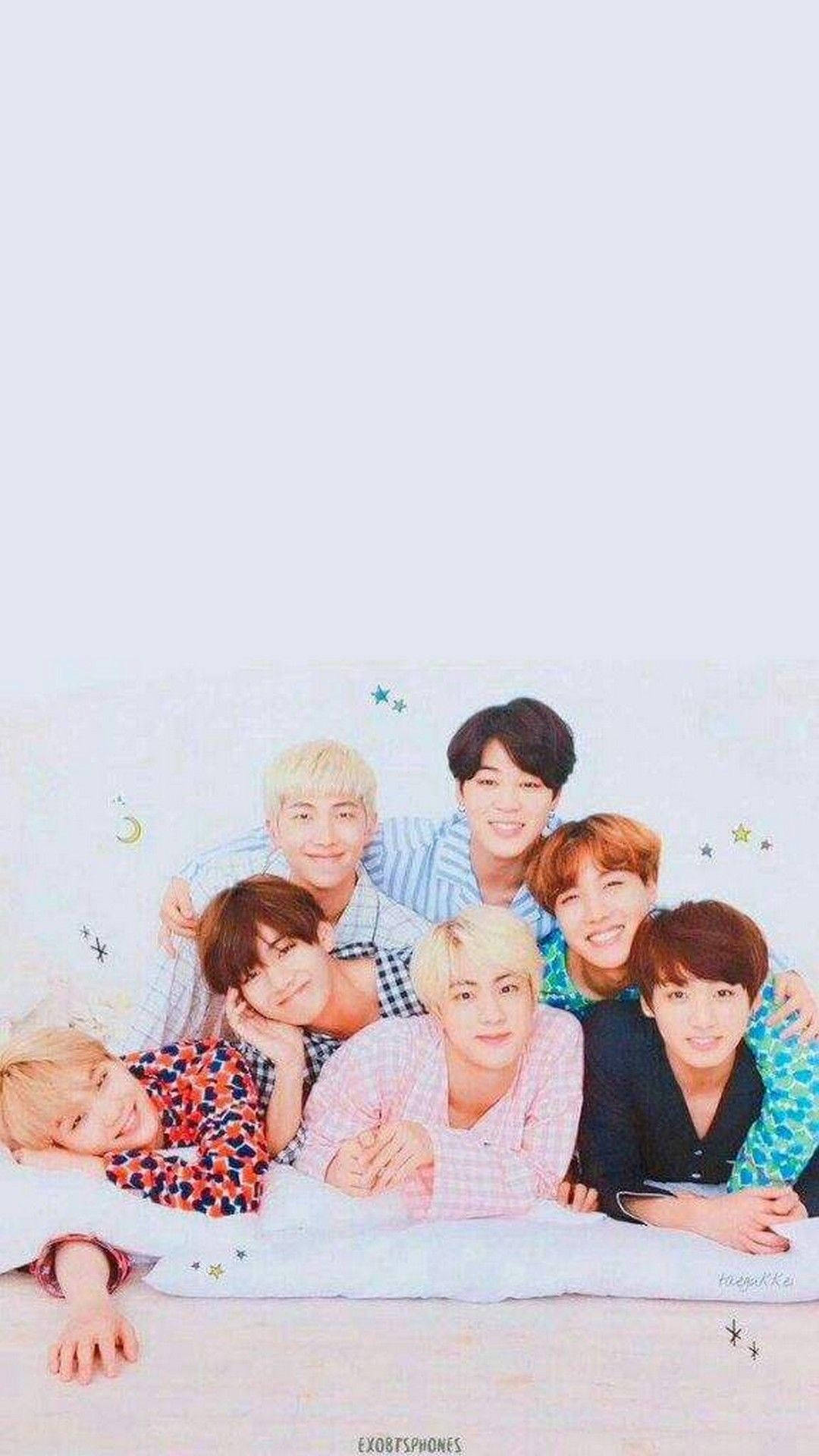 bts iPhone 桌布 壁紙 — 七位微笑的年輕男子身穿彩色圖案襯衫，在帶有星星和月亮裝飾的白色床上，以柔和粉彩背景一起擺姿勢。