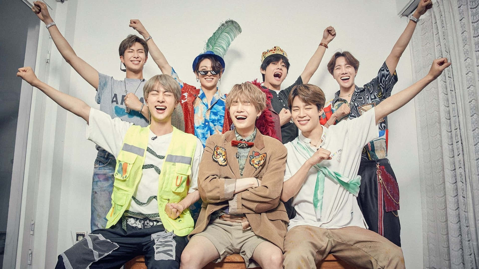 BTS 桌布 壁紙 — 七位微笑的年輕男子身穿色彩繽紛的服裝，在室內舉起雙臂，以素色牆壁和窗簾為背景，歡樂地擺姿勢。