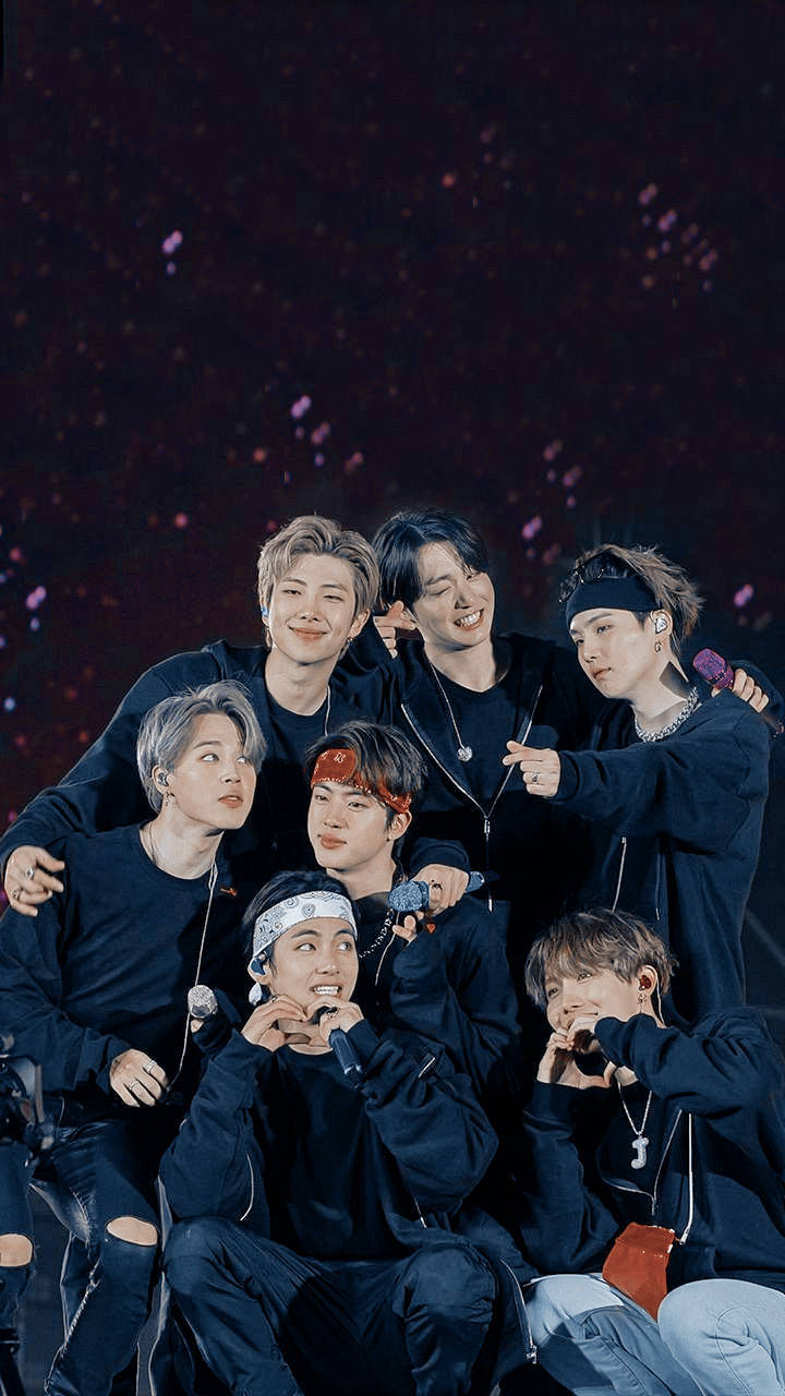 BTS 美學 桌布 壁紙 — 七位微笑的男性偶像穿著深色服裝在舞台上緊密擺姿勢，閃閃發光的紫色紙屑在他們周圍飄落。