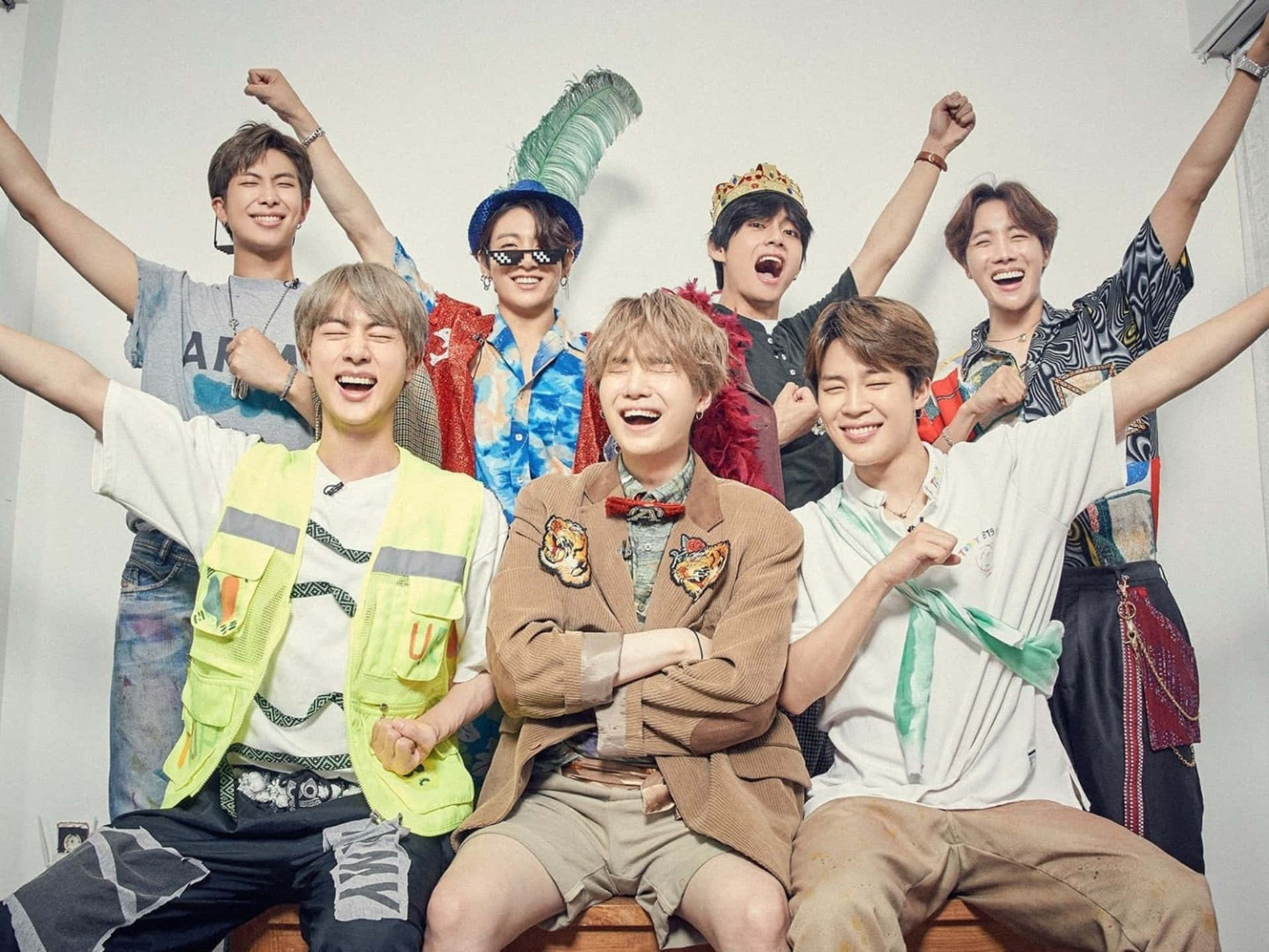 Bts Funny 桌布 壁紙 — 七位快樂的年輕男子穿著色彩繽紛、風格混搭的服裝，在純白牆壁前高舉雙臂擺姿勢。