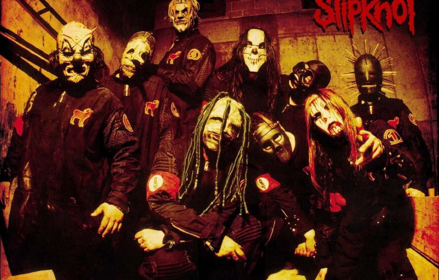 Slipknot 桌布 壁紙 — 七位戴面具的音樂家身穿黑色外套，在溫暖的橙色燈光下，在粗糙的磚牆前擺出強烈的姿勢。