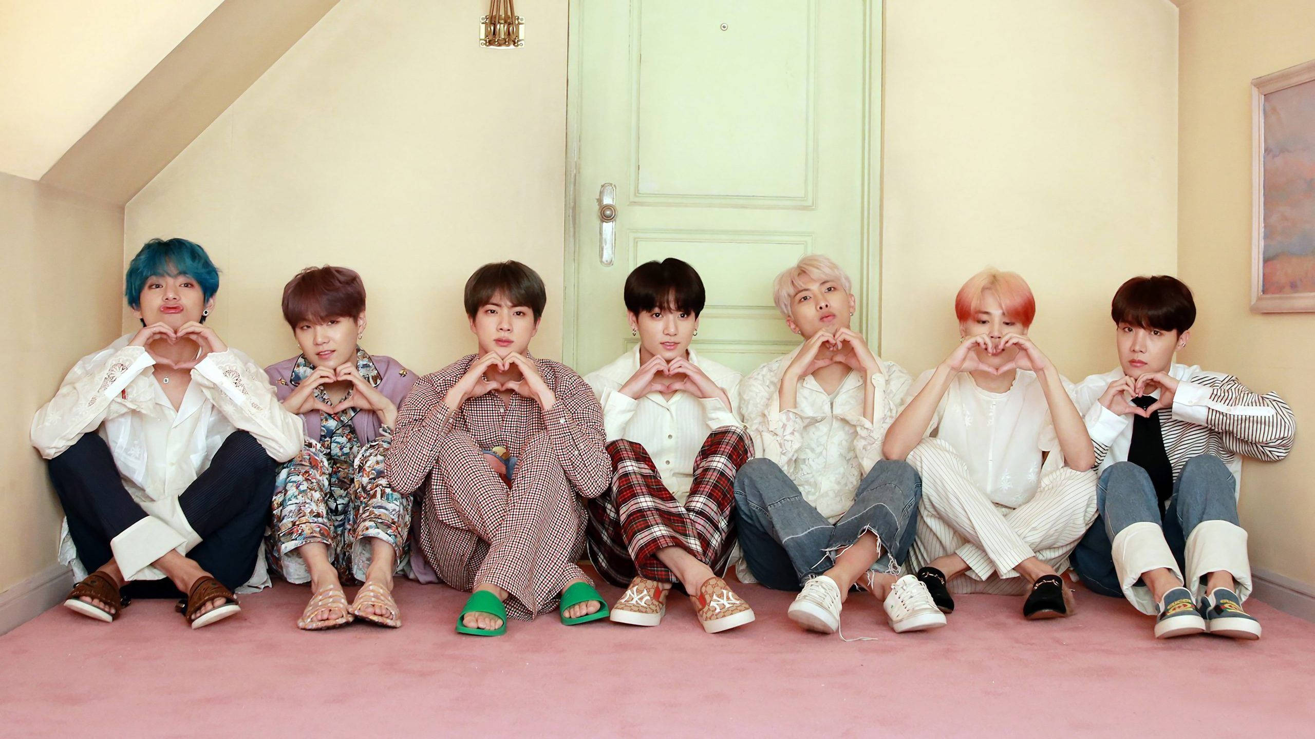 BTS 2021 桌布 壁紙 — 七位擁有彩色頭髮的年輕男性坐在傾斜閣樓房間的粉紅色地毯上，用手比出心形。