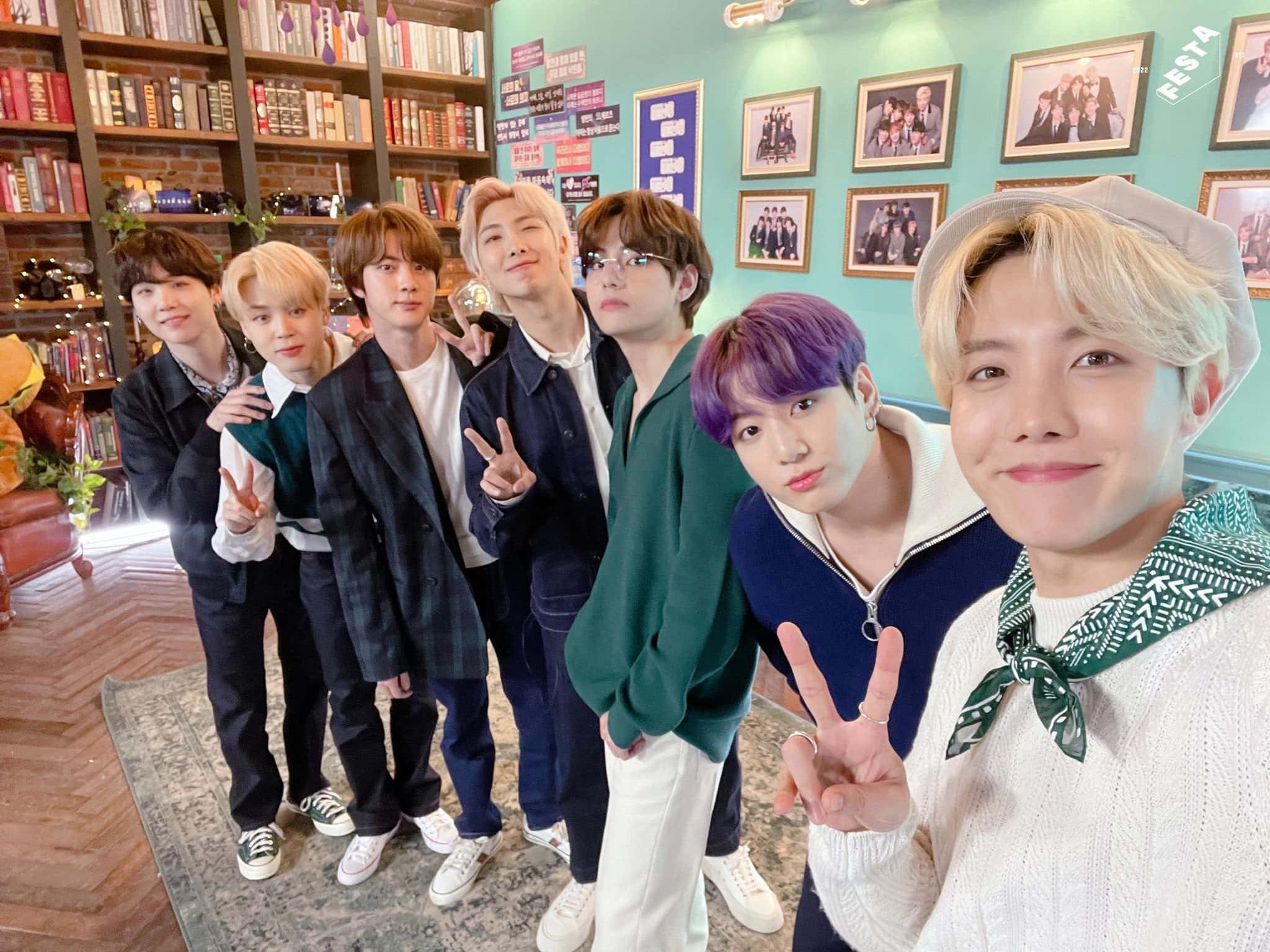 BTS 桌布 壁紙 — 七位擁有繽紛髮色的年輕男性在舒適的書店中愉快地擺姿勢，書店有書架和相框照片掛在青綠色的牆上。