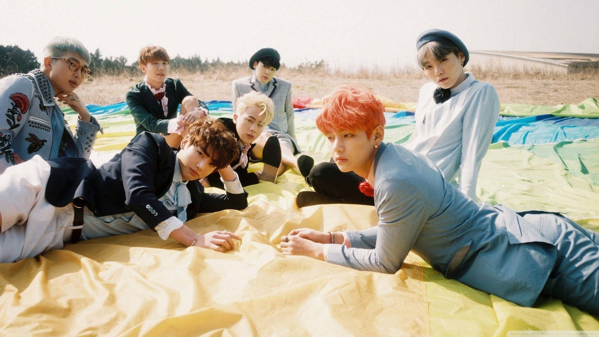 BTS 2021 桌布 壁紙 — 七位擁有色彩繽紛頭髮的年輕男子，在明亮的陽光下，於乾燥的草地上，躺在一張鮮黃色和藍色的防水布上休憩。