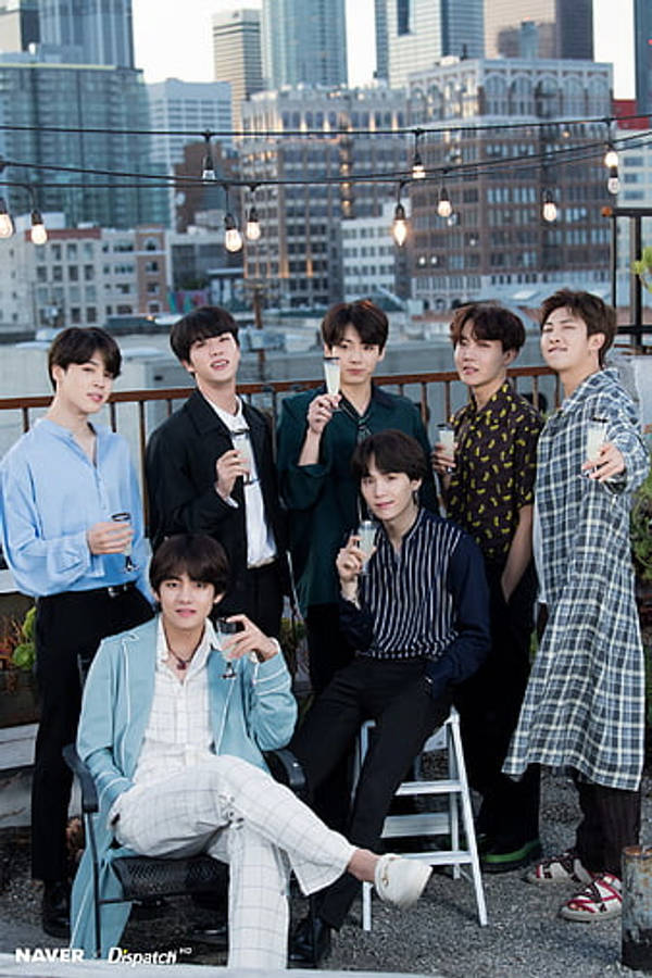 BTS 桌布 壁紙 — 七位時尚年輕男士在屋頂露臺上隨意擺姿勢，背景是城市天際線和燈串。