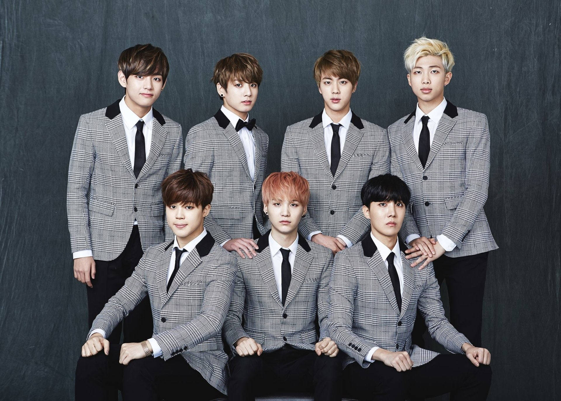BTS 桌布 壁紙 — 七位時尚年輕男士身穿相配的灰色格紋西裝，在深色紋理背景前自信地擺姿勢。