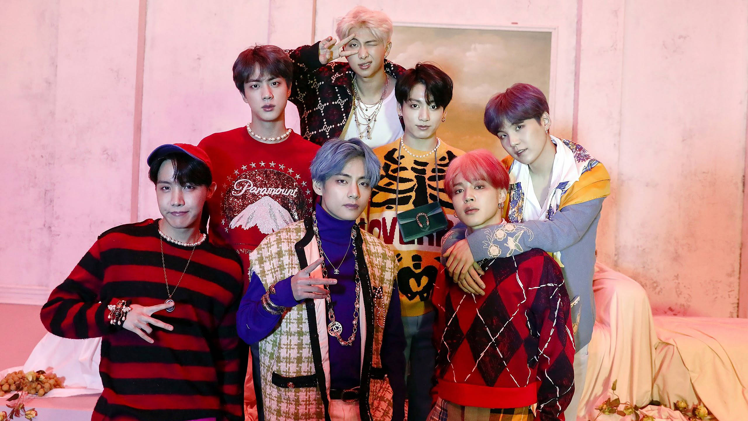 BTS 桌布 壁紙 — 七位時尚年輕男士身穿色彩繽紛的圖案服裝，在帶有花卉裝飾的柔軟粉紅色紋理牆前自信地擺姿勢。