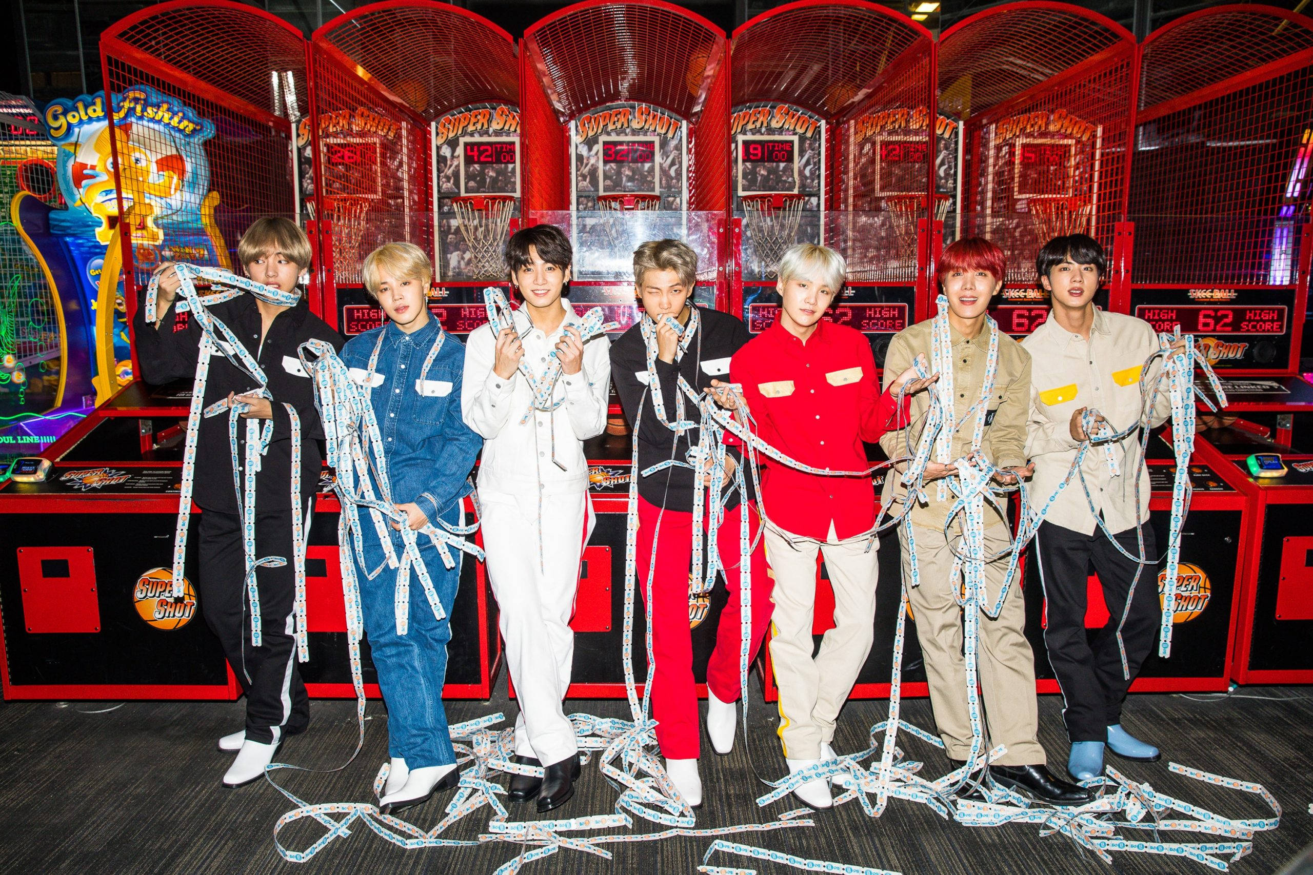 BTS 桌布 壁紙 — 七位時尚年輕男子在充滿活力的遊戲機中心自信地擺姿勢，手持纏結的漁網，背景是紅色遊戲機。