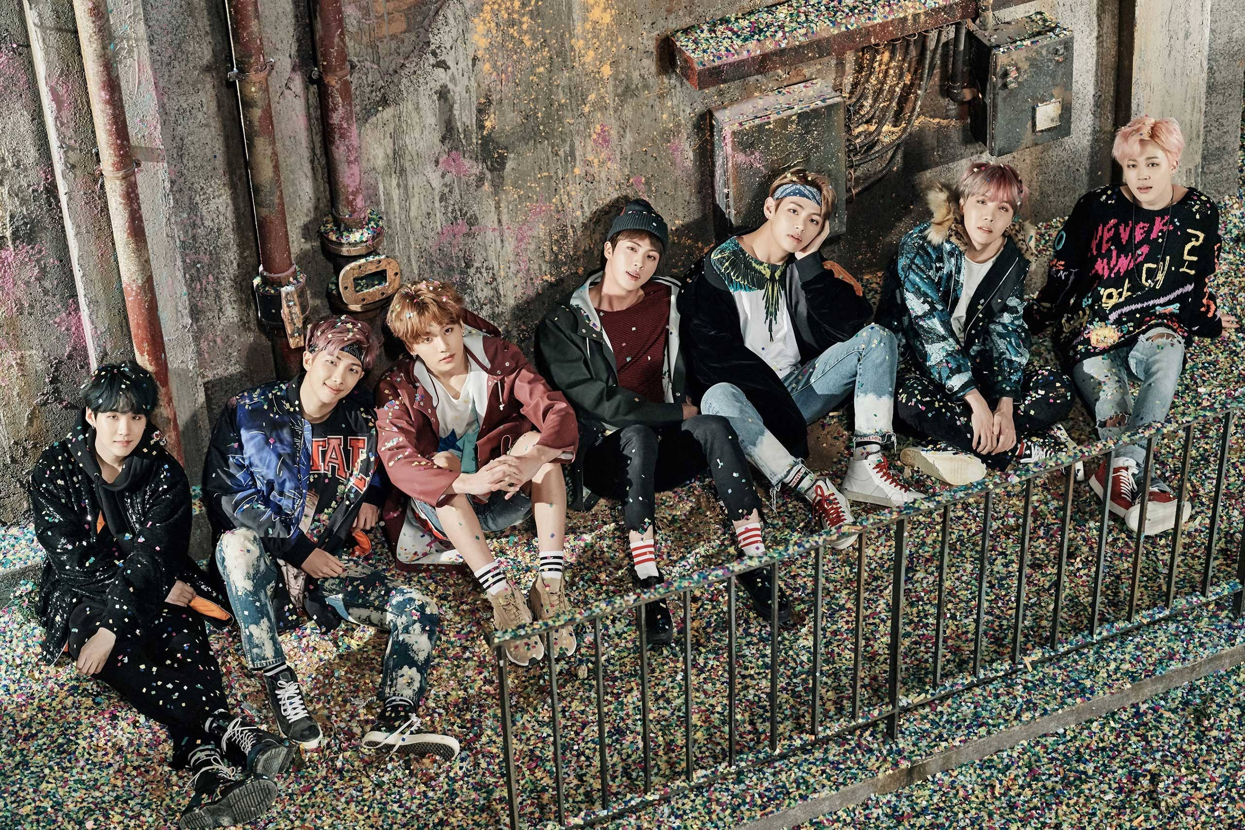 BTS 筆電 桌布 壁紙 — 七位時尚年輕男子自信地站在金屬圍欄上，身處佈滿彩色紙屑的工業小巷中。