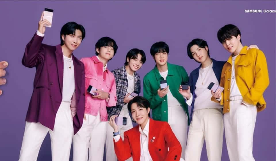 Bts Endorsements 桌布 壁紙 — 七位時尚年輕男子身穿彩色外套，在純紫色背景前愉快地擺姿勢，每人手持一部智慧型手機。