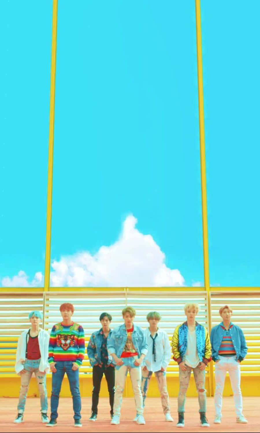 Bts Mv 桌布 壁紙 — 七位時尚年輕男子身穿色彩繽紛的服裝，自信地站在由黃色垂直樑柱框住的明亮藍天下。
