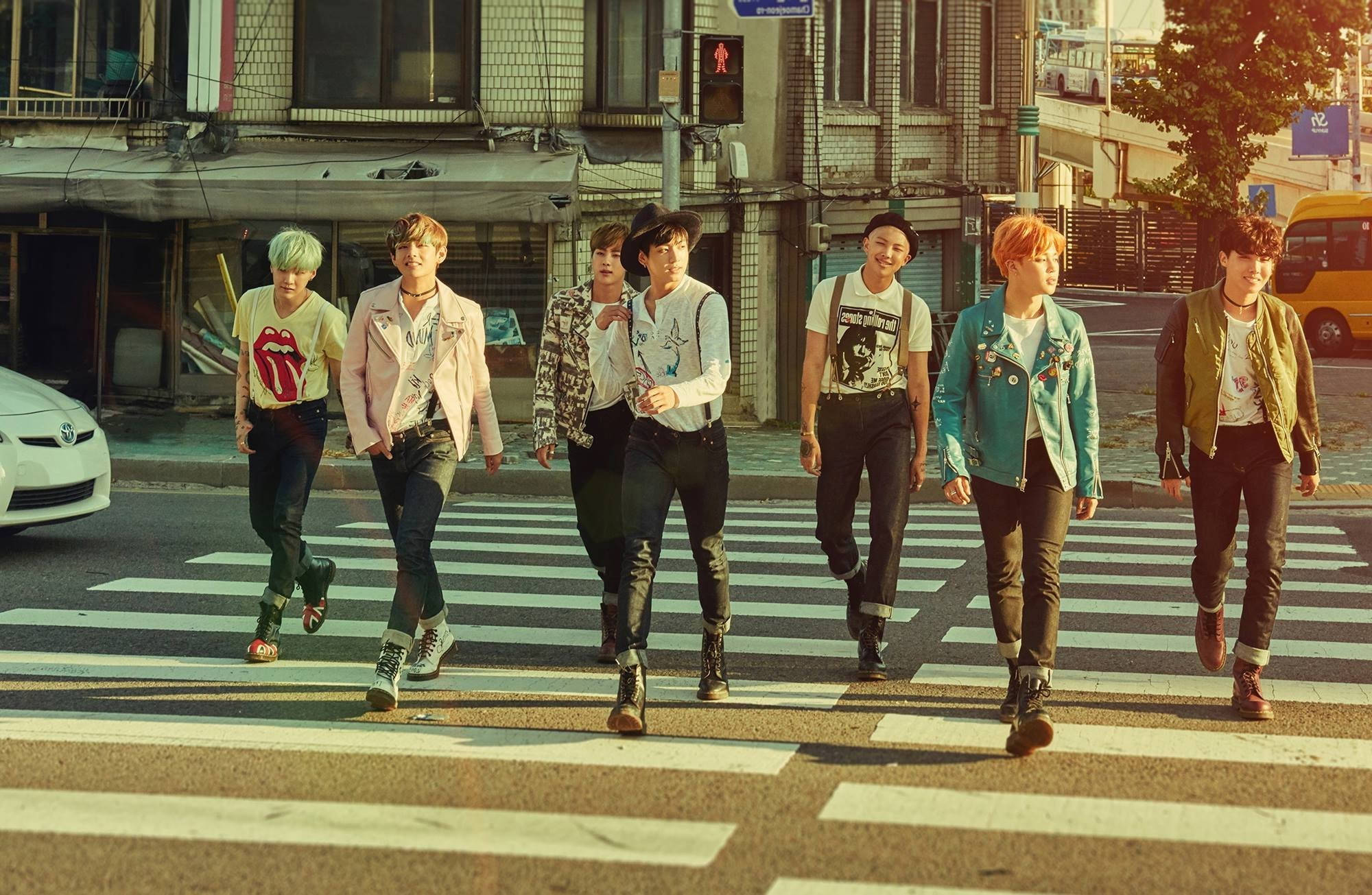 BTS 桌布 壁紙 — 七位時尚年輕男子身穿色彩繽紛的街頭服飾，自信地大步走過陽光普照的城市行人穿越道，背景有建築物和汽車。