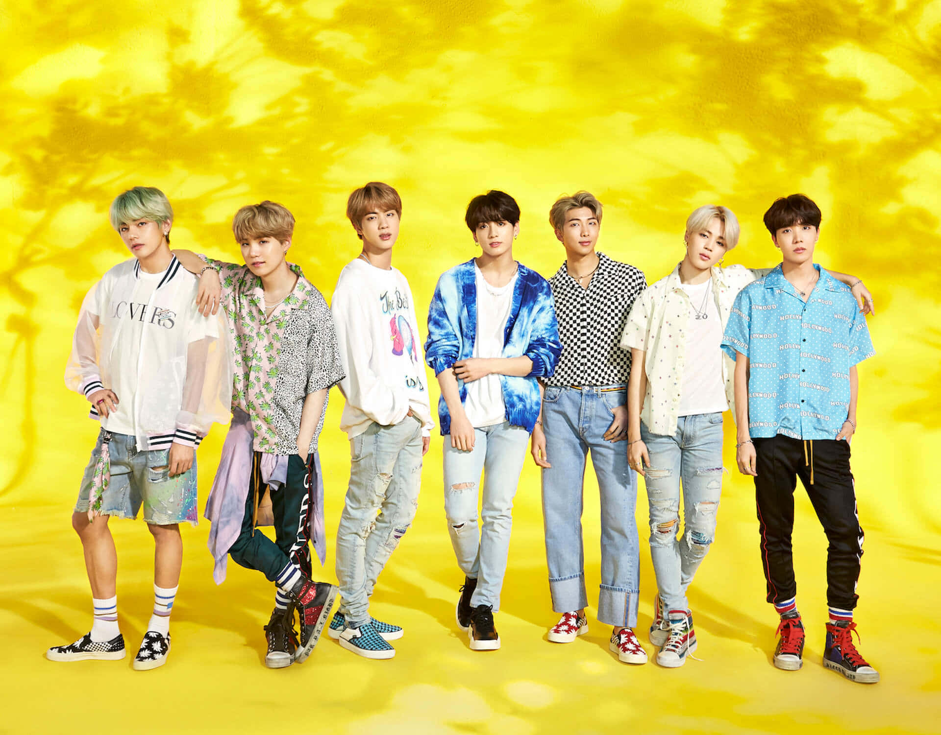 Bts Mv 桌布 壁紙 — 七位時尚年輕男子身穿色彩繽紛的街頭服飾，在充滿活力的黃色背景前自信地擺姿勢，背景有棕櫚樹陰影。