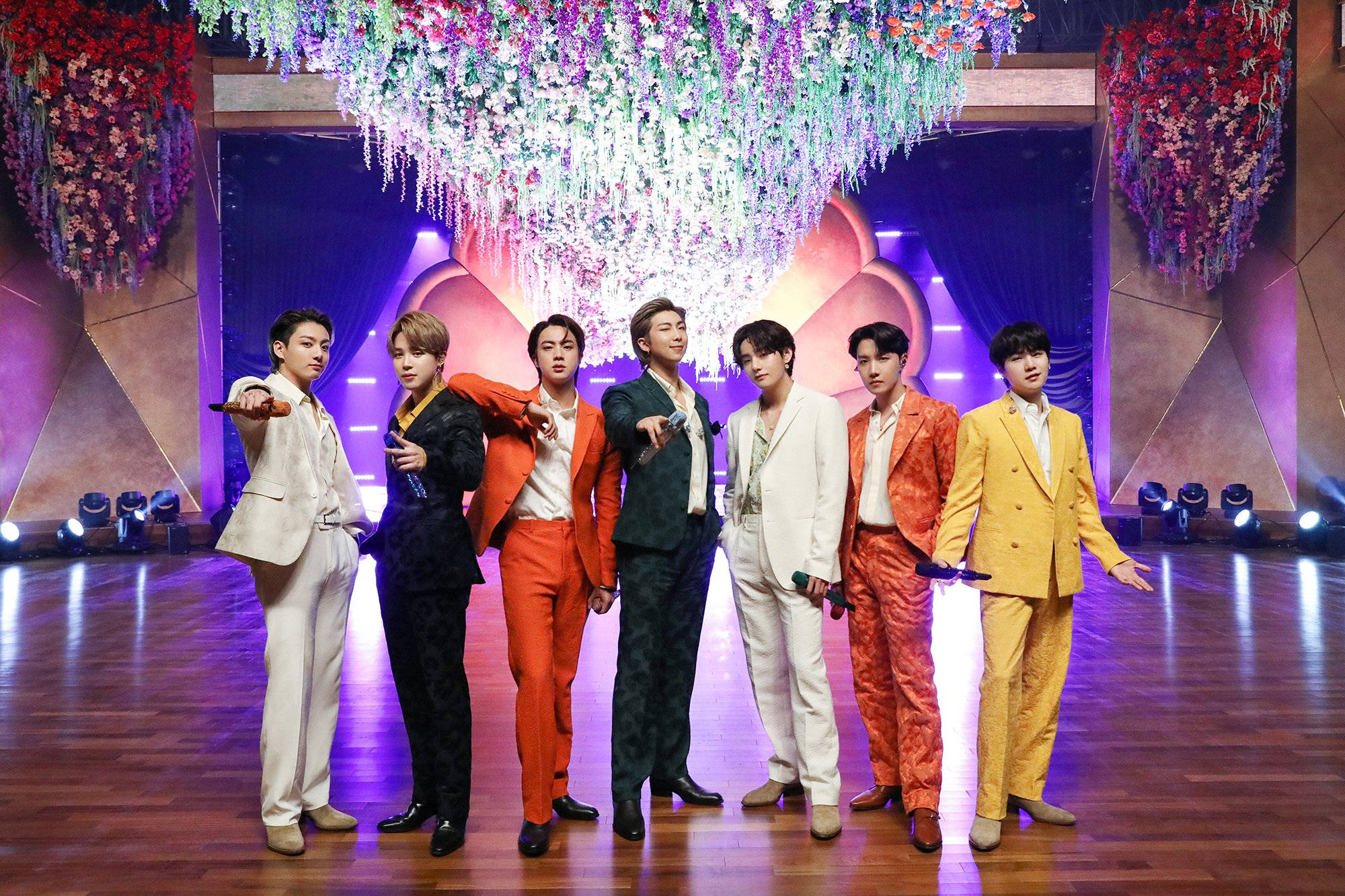 BTS 2021 桌布 壁紙 — 七位時尚男士穿著大膽色彩的西裝，在充滿活力的舞台上，於層疊的花卉吊燈下自信地擺姿勢。