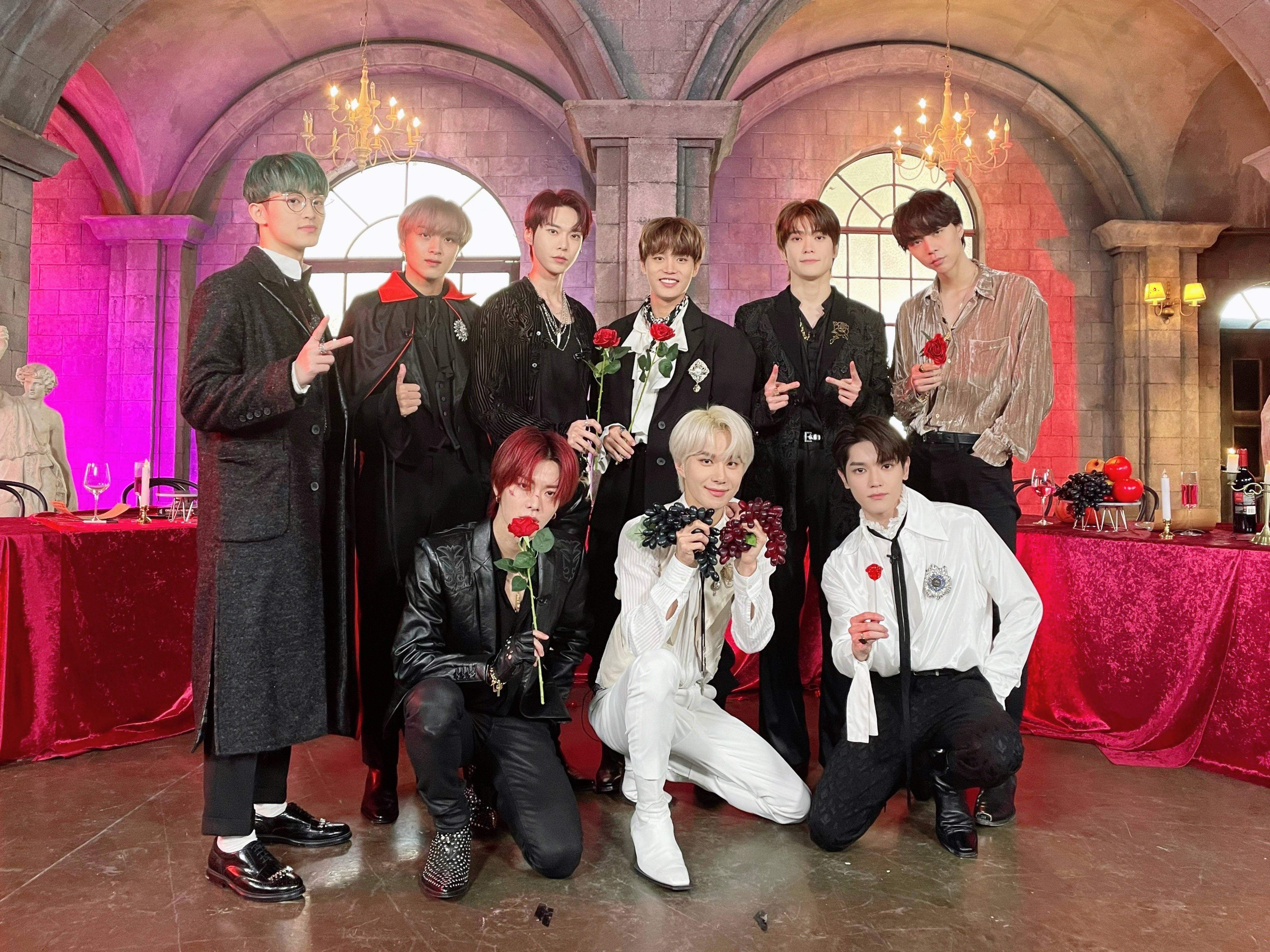 Nct 桌布 壁紙 — 七位時尚男士穿著正式服裝，在一個宏偉的哥德式大廳中，與紅玫瑰和葡萄一起擺姿勢，大廳內有吊燈和紅色天鵝絨桌子。