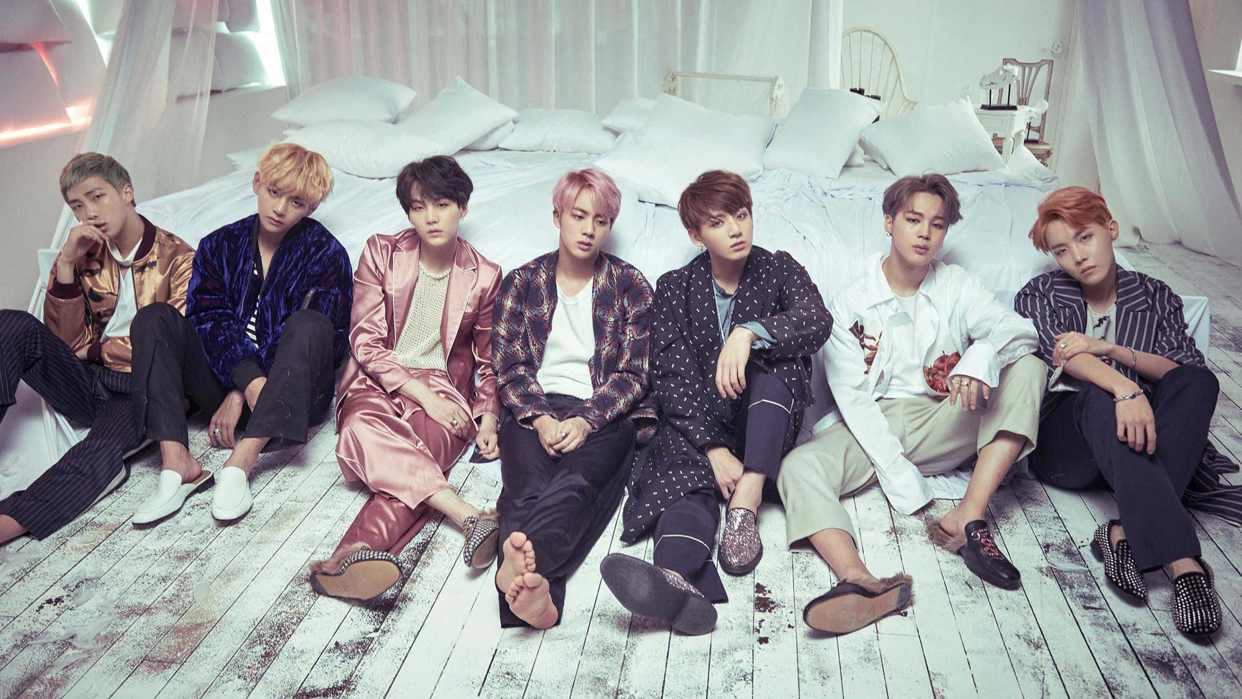 Bts Mac 桌布 壁紙 — 七位時尚男士穿著色彩繽紛的服裝，隨意地坐在一個光線充足、通風良好、鋪有木地板的房間裡的白色床上。