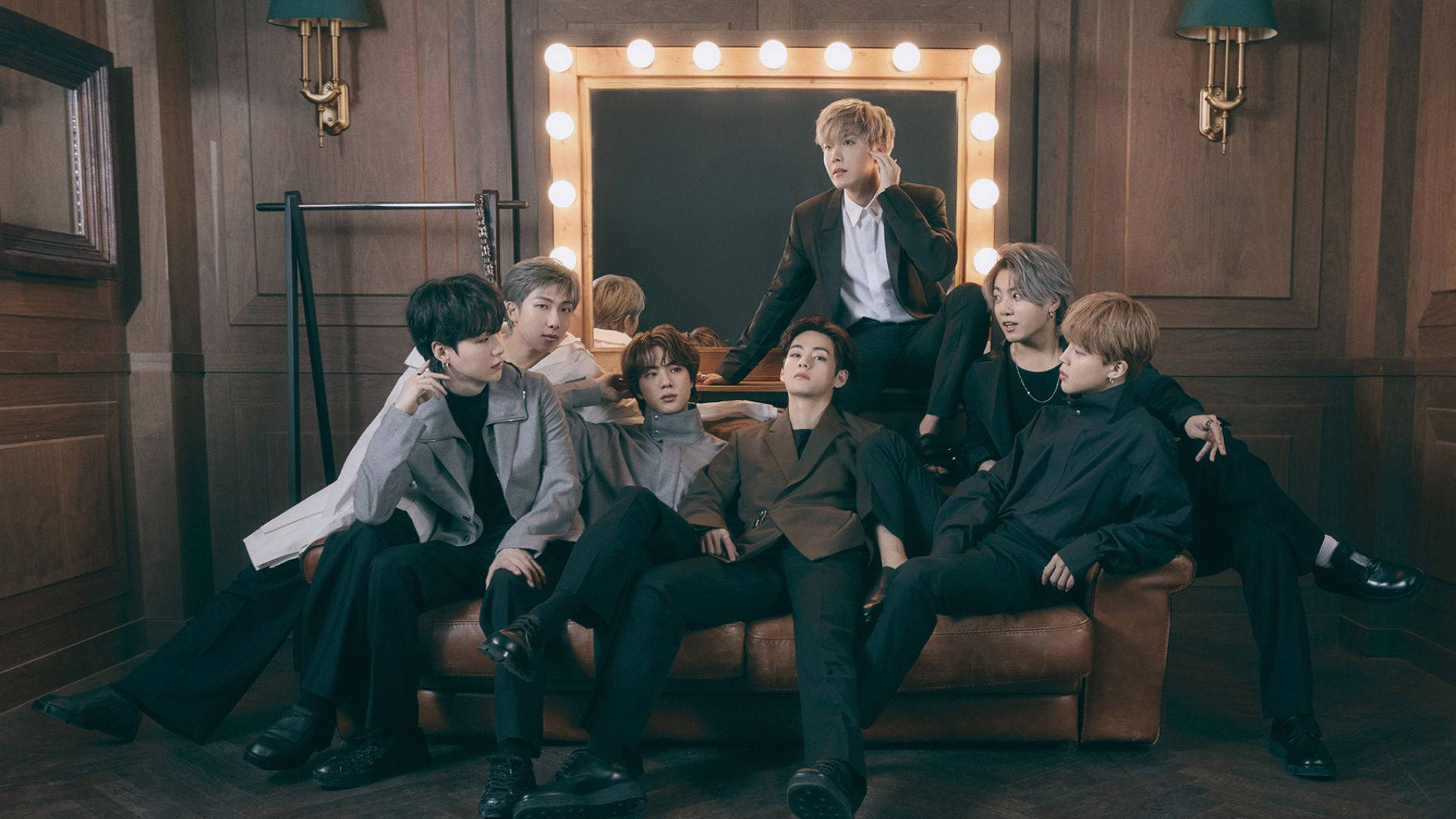 BTS 桌布 壁紙 — 七位時尚男士自信地坐在一張皮革沙發上，身處一個光線溫暖的木製房間，旁邊有一面化妝鏡。