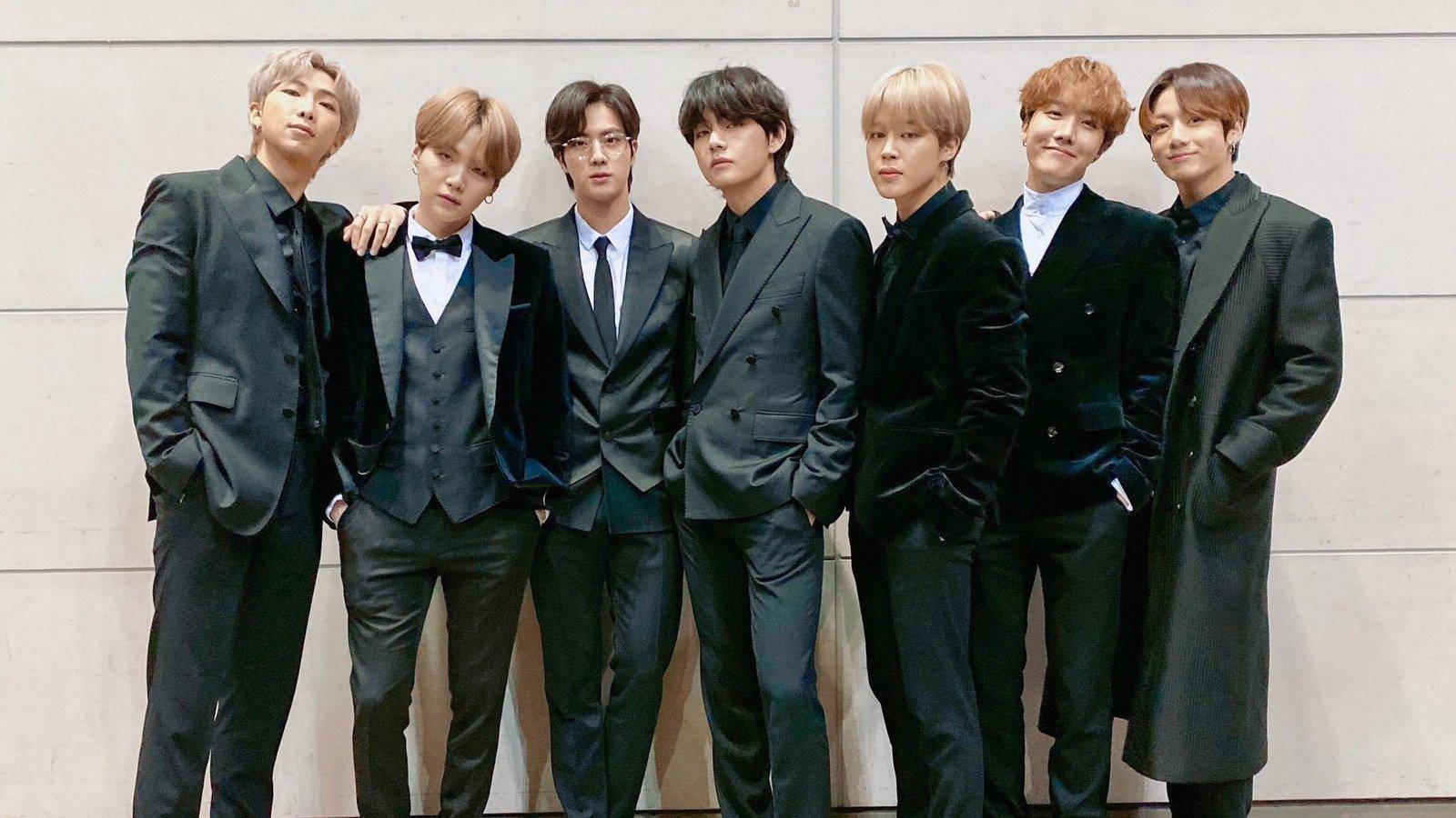 BTS 桌布 壁紙 — 七位時尚男士身穿俐落黑色西裝，以多樣髮型和表情，自信地站在簡約米色牆前擺姿勢。