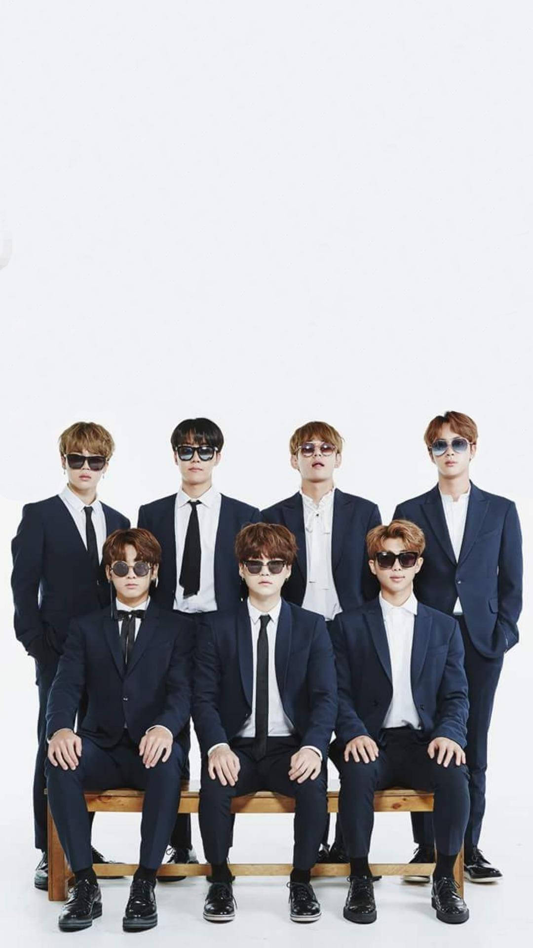 BTS 桌布 壁紙 — 七位時尚男士身穿相配的海軍藍西裝和太陽眼鏡，在白色背景下的木長椅上自信地擺姿勢。