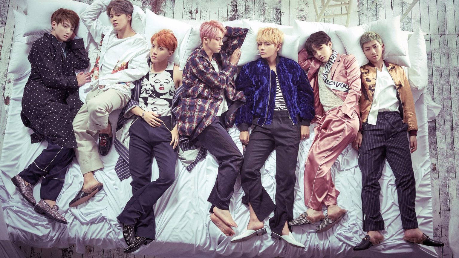 BTS 桌布 壁紙 — 七位時尚男士身穿色彩繽紛的服裝，在白色床上靠著枕頭，背景是風化的木牆。