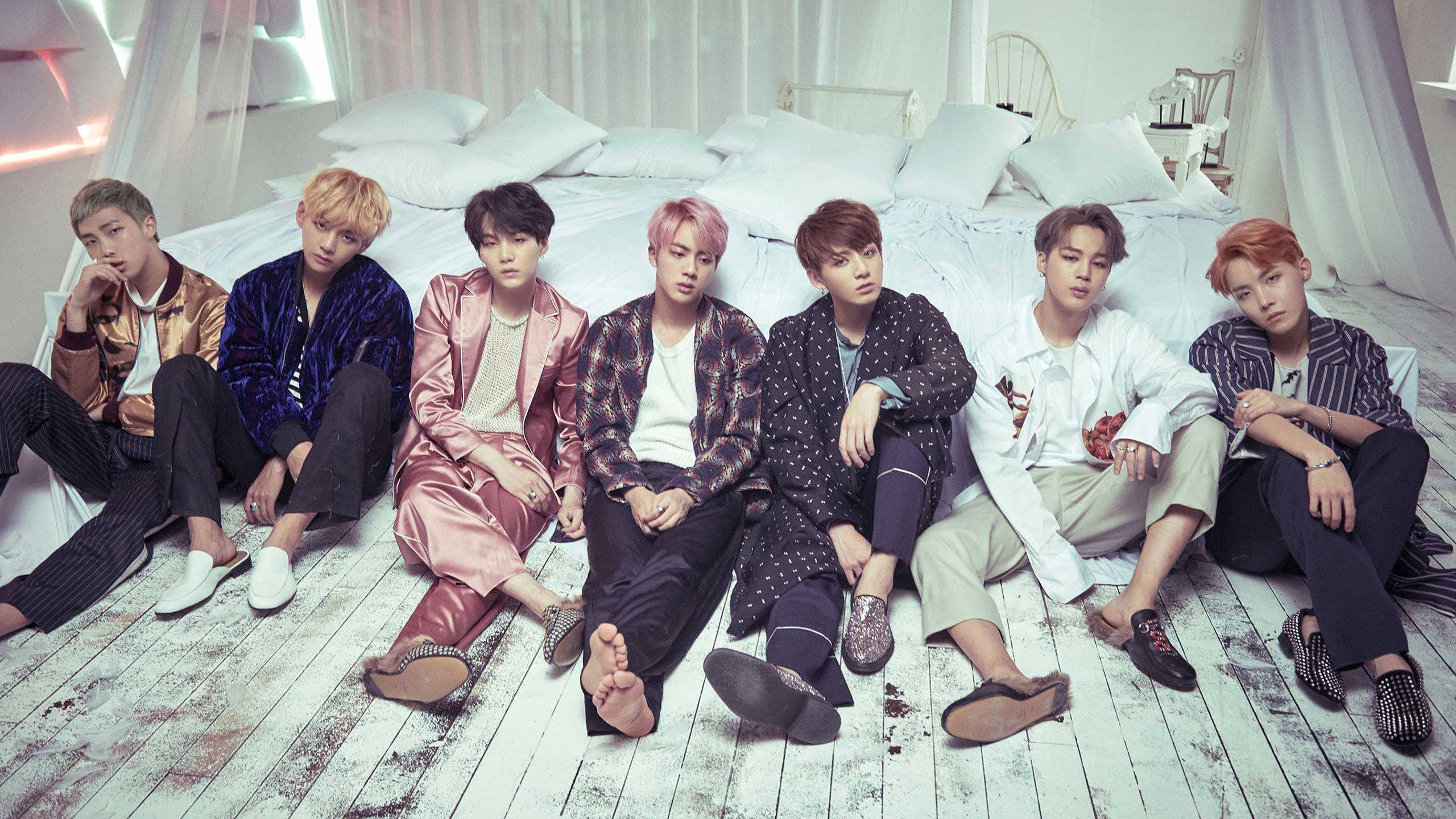 BTS 桌布 壁紙 — 七位時尚男士身穿色彩繽紛的服裝，隨意地坐在白色木地板上，旁邊是一張配有白色枕頭和薄紗窗簾的床。