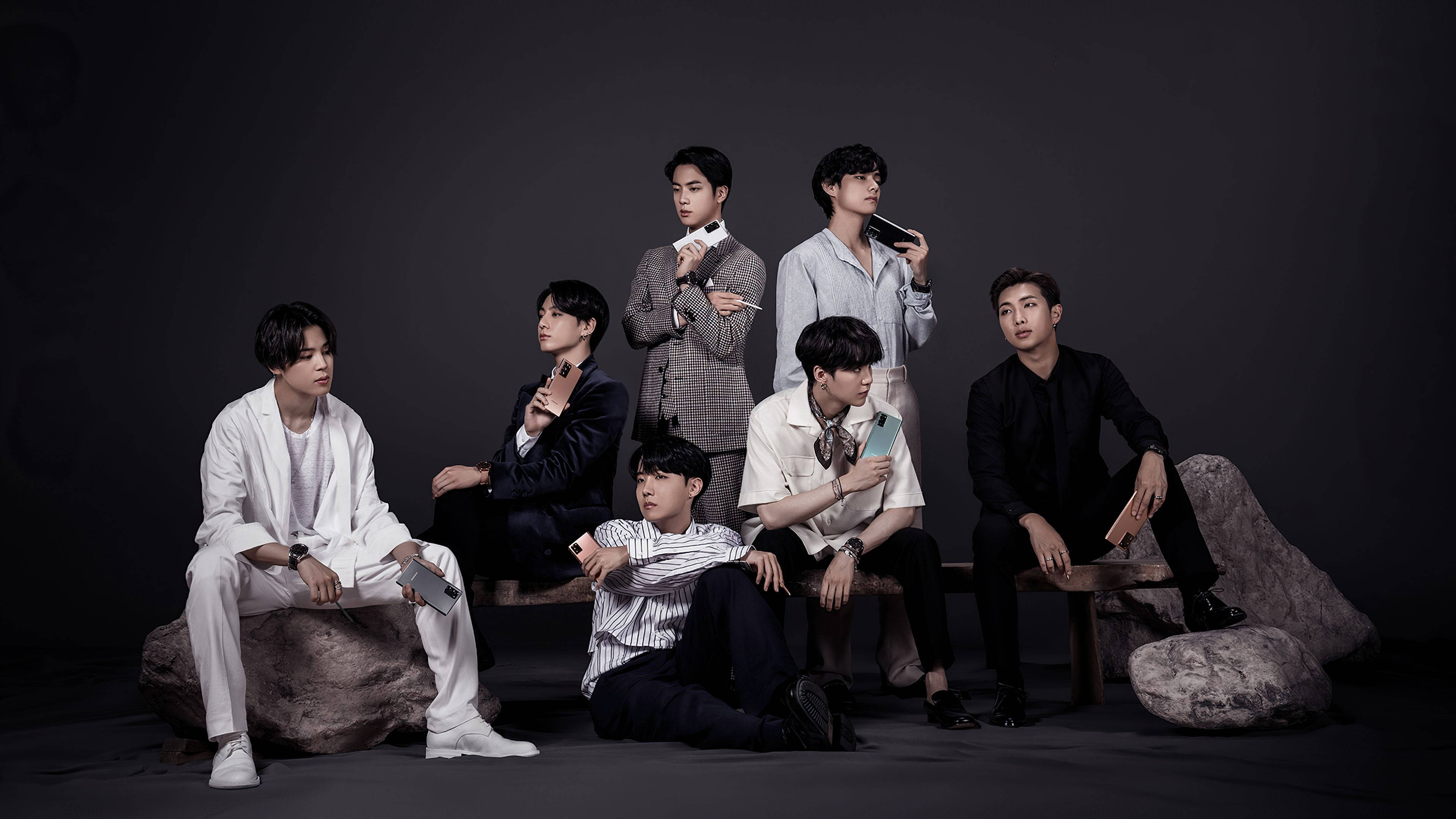 BTS 桌布 壁紙 — 七位時尚男士身著現代服裝，在黑暗的攝影棚背景下隨意地坐在岩石和長椅上擺姿勢。