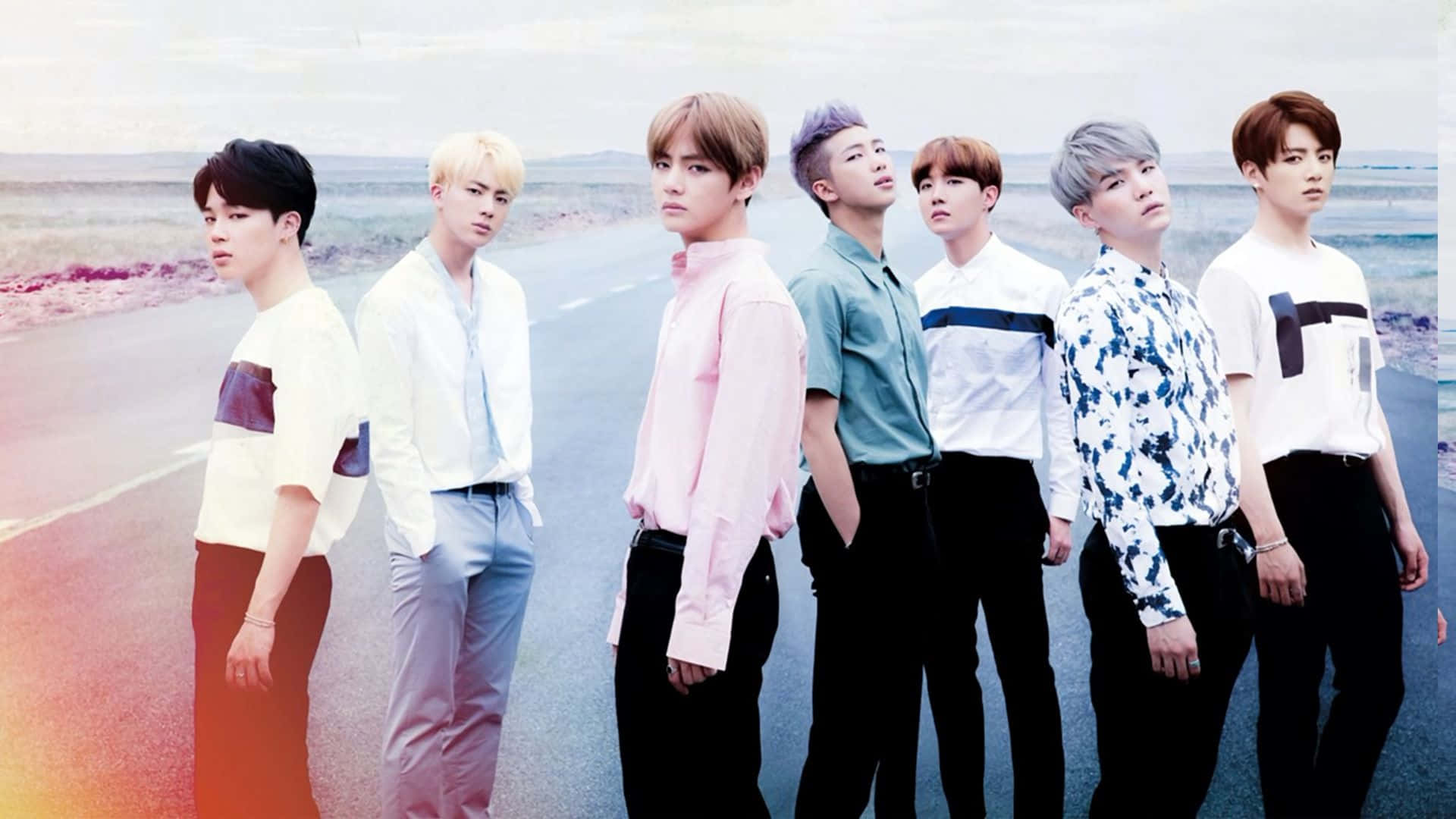 K-pop 桌布 壁紙 — 七位時尚的年輕男士身穿色彩繽紛的襯衫，自信地站在沙漠公路上，天空柔和。