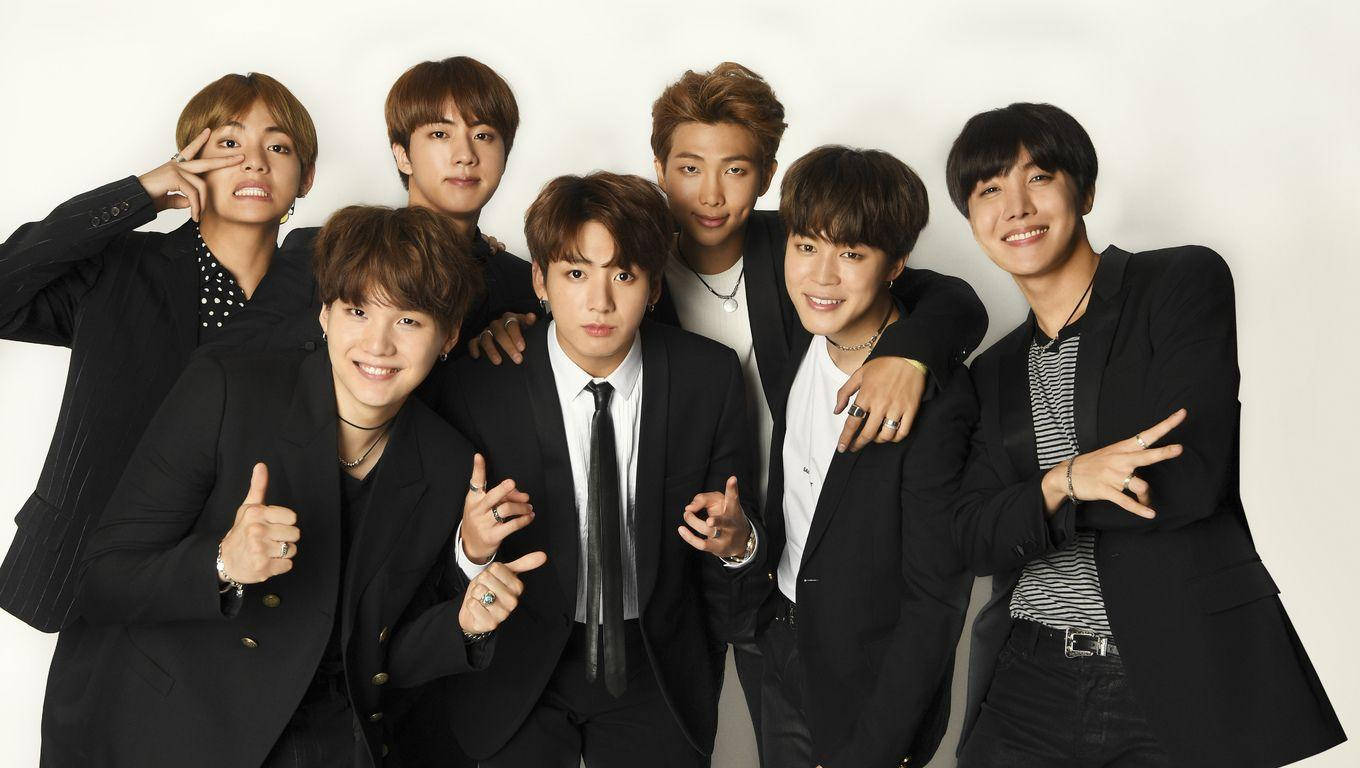 BTS 桌布 壁紙 — 七位時尚的年輕男士身穿黑色西裝，在純白色背景前自信地擺出俏皮的手勢。