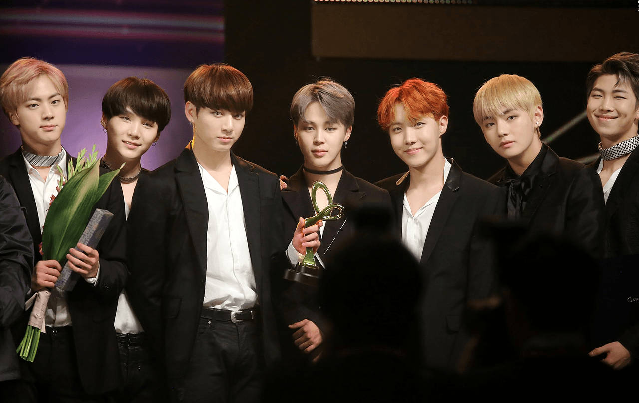 BTS 桌布 壁紙 — 七位時尚的年輕男士身穿黑色西裝，在明亮的燈光下自信地在舞台上擺姿勢，手持獎盃和花束。