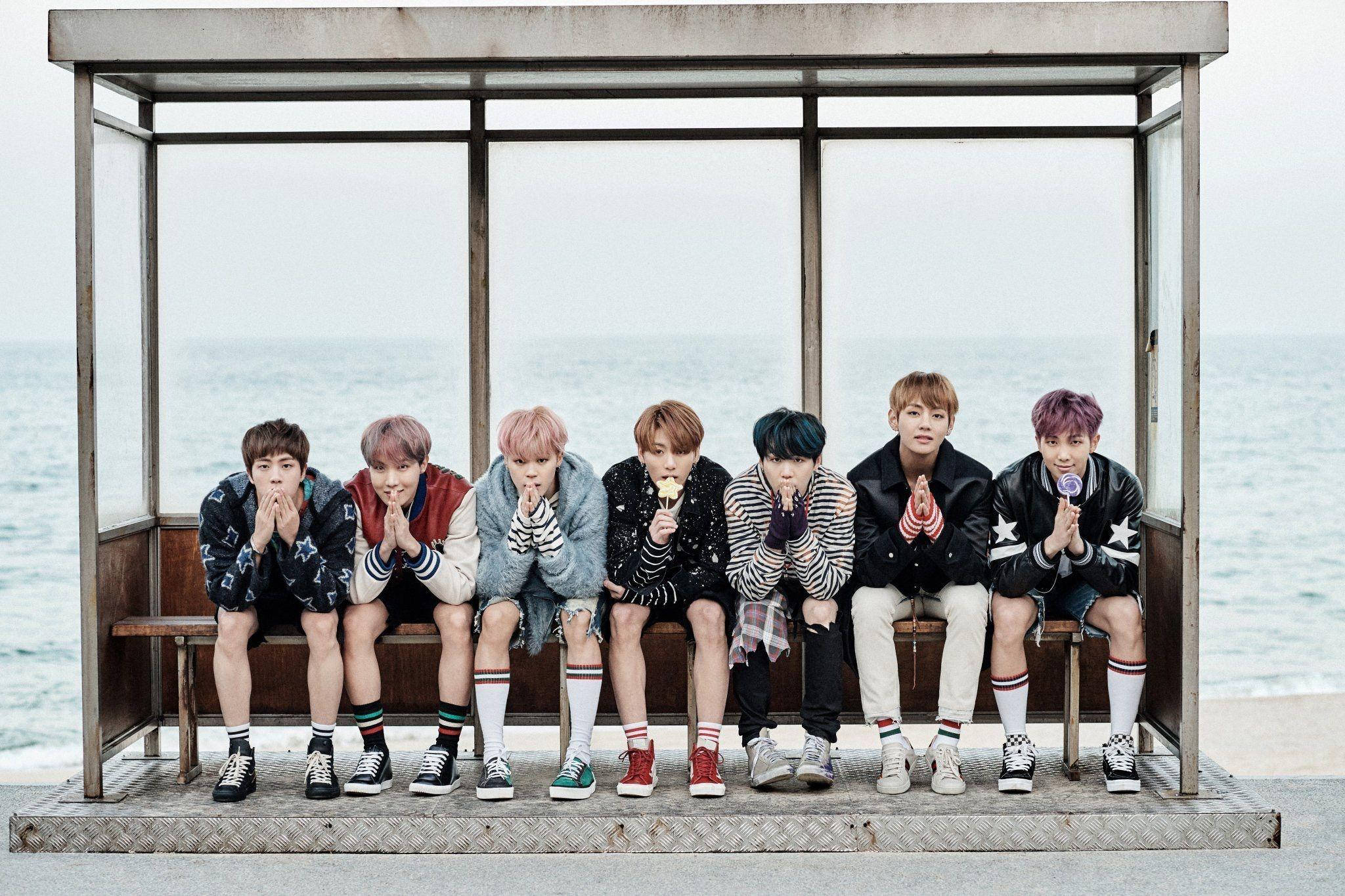 BTS 桌布 壁紙 — 七位時尚的年輕男子，擁有色彩繽紛的頭髮，坐在海邊公車站的長椅上，每人手持一支棒棒糖，背景是柔和的海景。