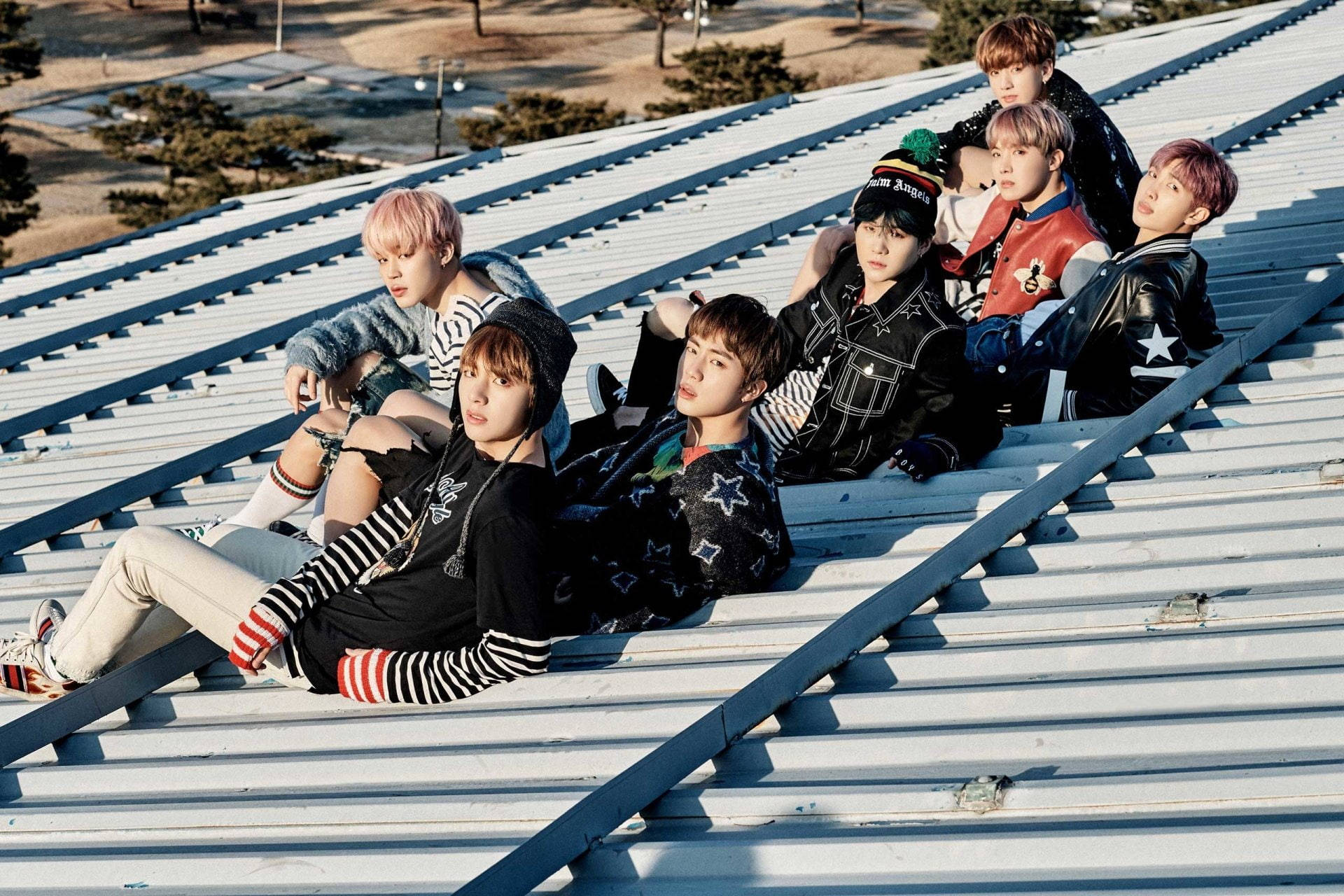 BTS 桌布 壁紙 — 七位時尚的年輕男子，頭髮色彩繽紛，穿著搭配的街頭服飾，在陽光普照的金屬屋頂上悠閒地休息。