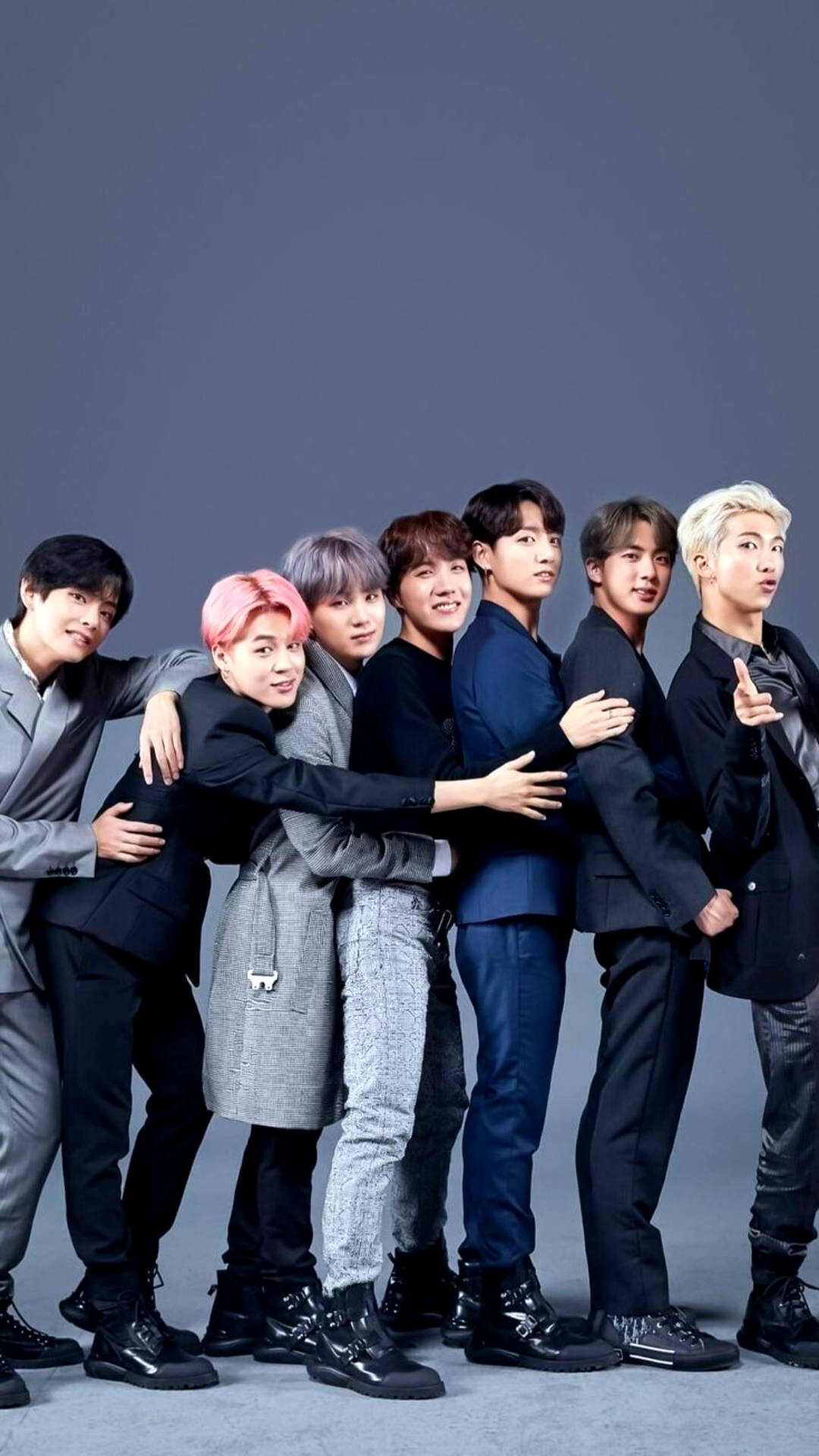BTS 鎖定螢幕 桌布 壁紙 — 七位時尚的年輕男子身穿深色西裝，頭髮色彩繽紛，在純灰色攝影棚背景前緊密地擺姿勢。