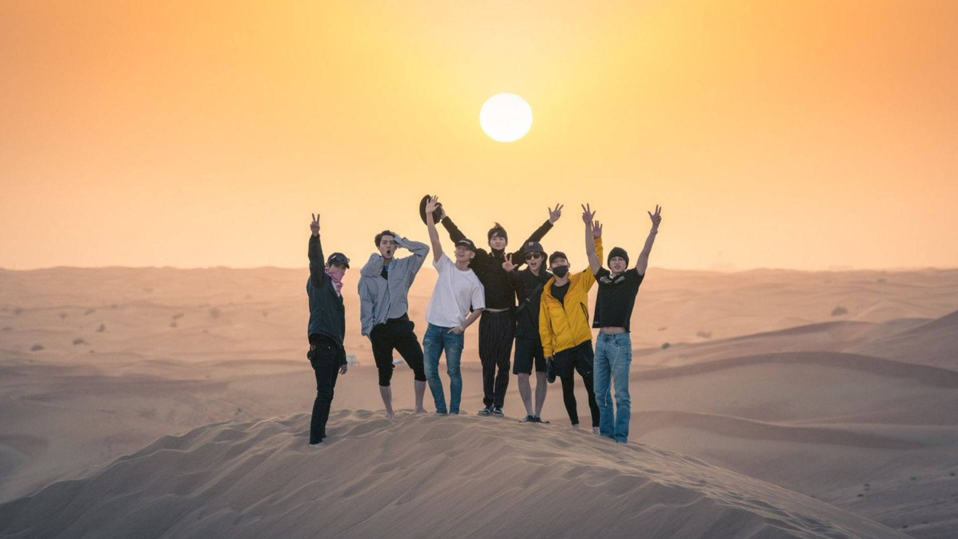 EXO 桌布 壁紙 — 七位朋友在沙丘頂上歡快地舉起雙臂，夕陽在溫暖的金色天空中落下。