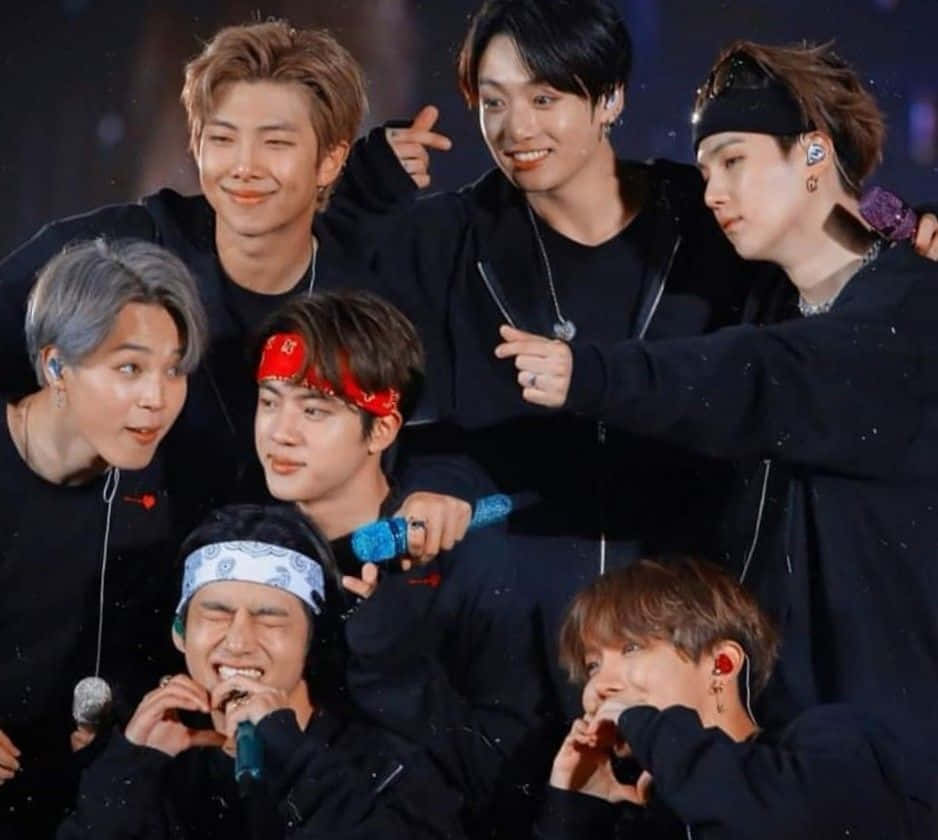 Jungkook 桌布 壁紙 — 七位男K-pop偶像身穿黑色服裝，在明亮的燈光下，手持麥克風和彩色頭帶，在舞台上俏皮地擺姿勢。