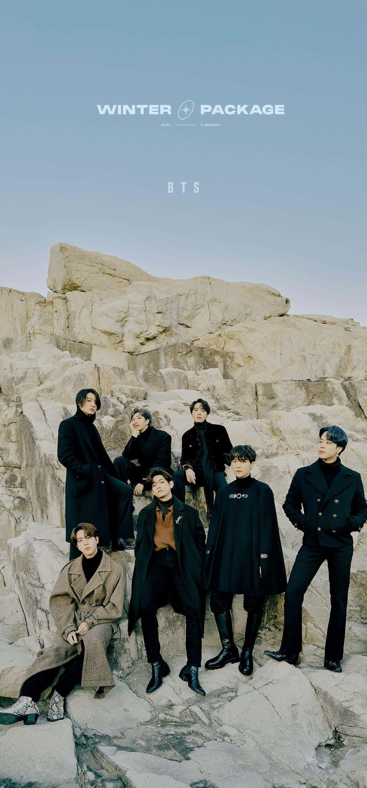 K-pop 桌布 壁紙 — 七位男士身穿時尚深色大衣，在淡藍色天空下，自信地站在崎嶇的岩石懸崖上。