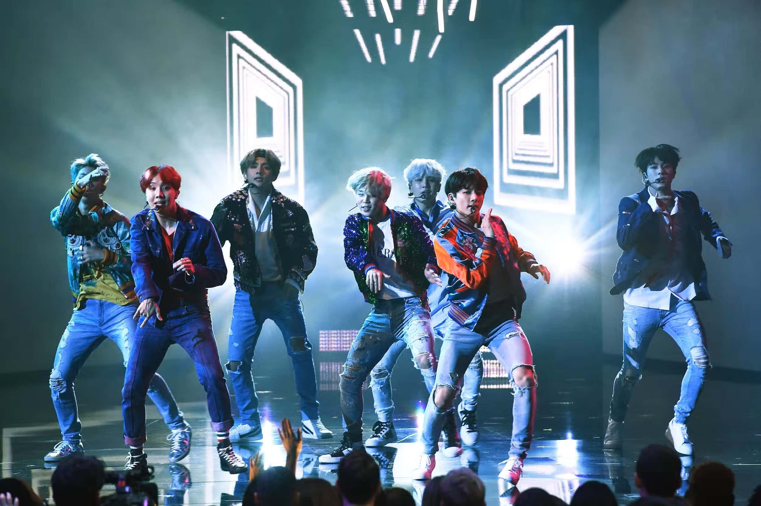 Bts Performance 桌布 壁紙 — 七位男性表演者身穿彩色外套和破洞牛仔褲，在明亮的舞台燈光下，以幾何背景為襯，充滿活力地跳舞。