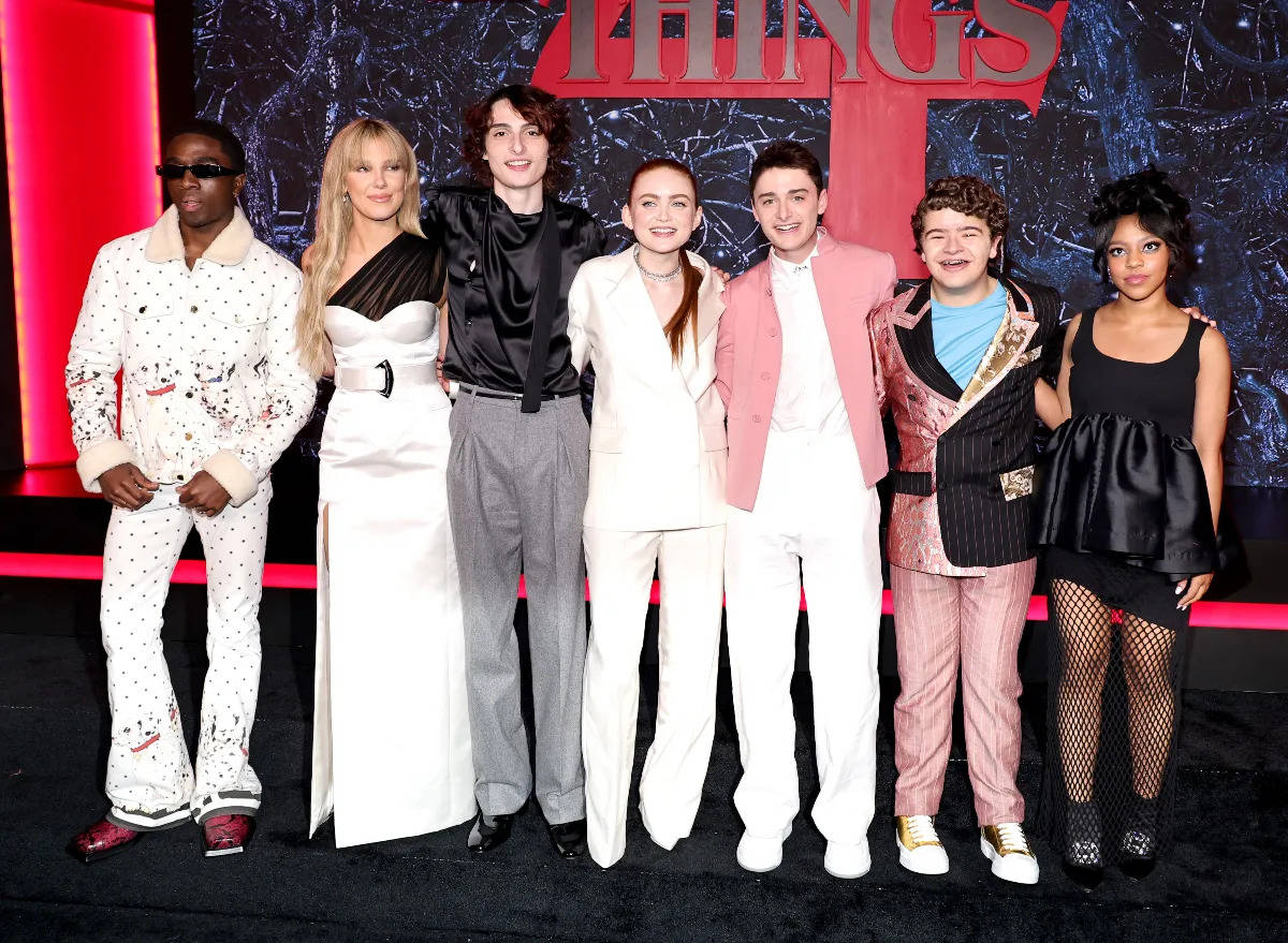 Stranger Things 4 桌布 壁紙 — 七位穿著時尚的名人在紅地毯上自信地擺姿勢，背景是醒目的紅黑色背景，上面有「THINGS」文字。